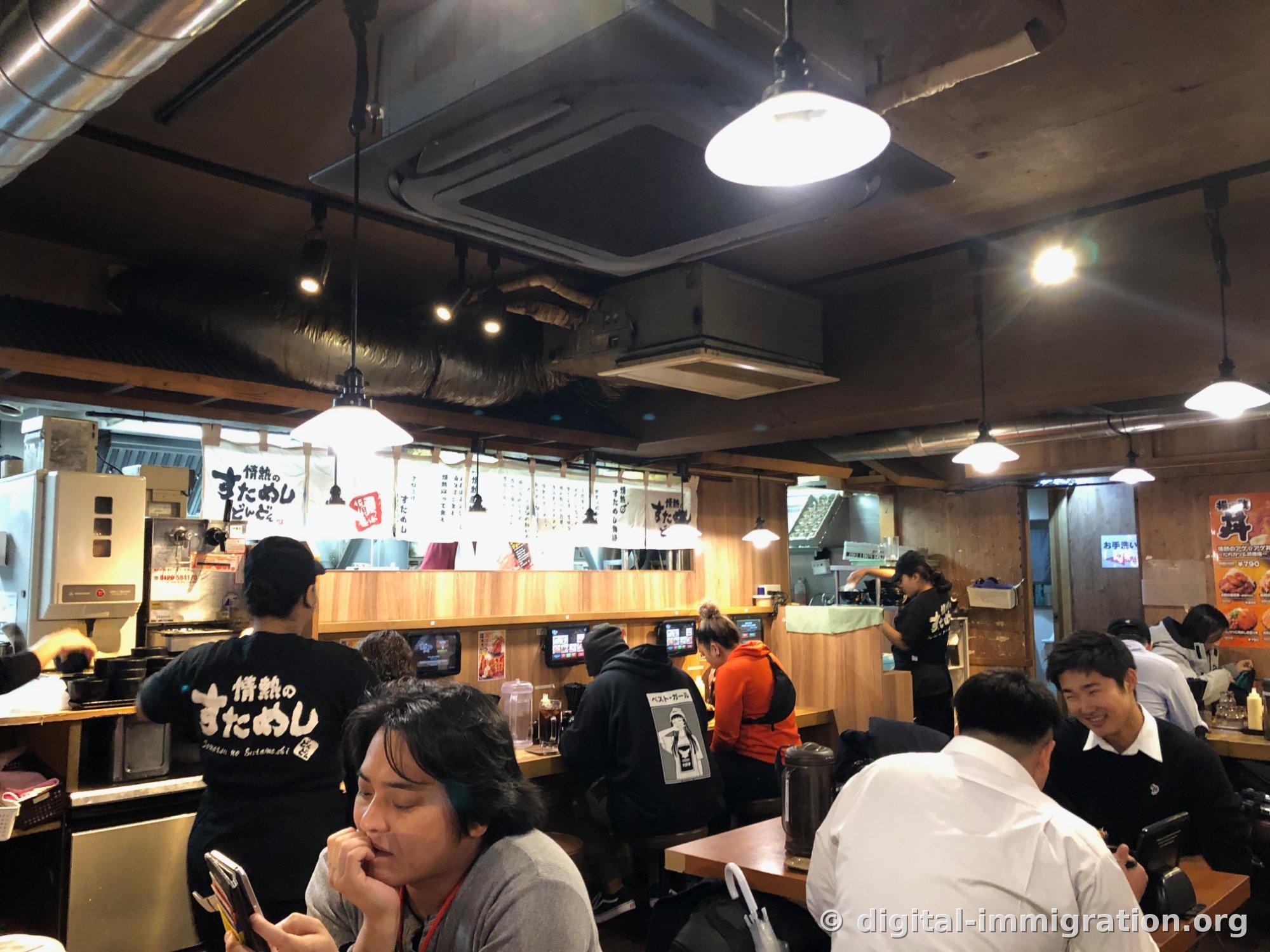 Intérieur du hall souterrain, avec les tuyauteries exposées au plafond et les luminaires suspendus, les clients assis aux places de comptoir, et les employés en uniforme noir avec le caractère すためし imprimé sur le dos