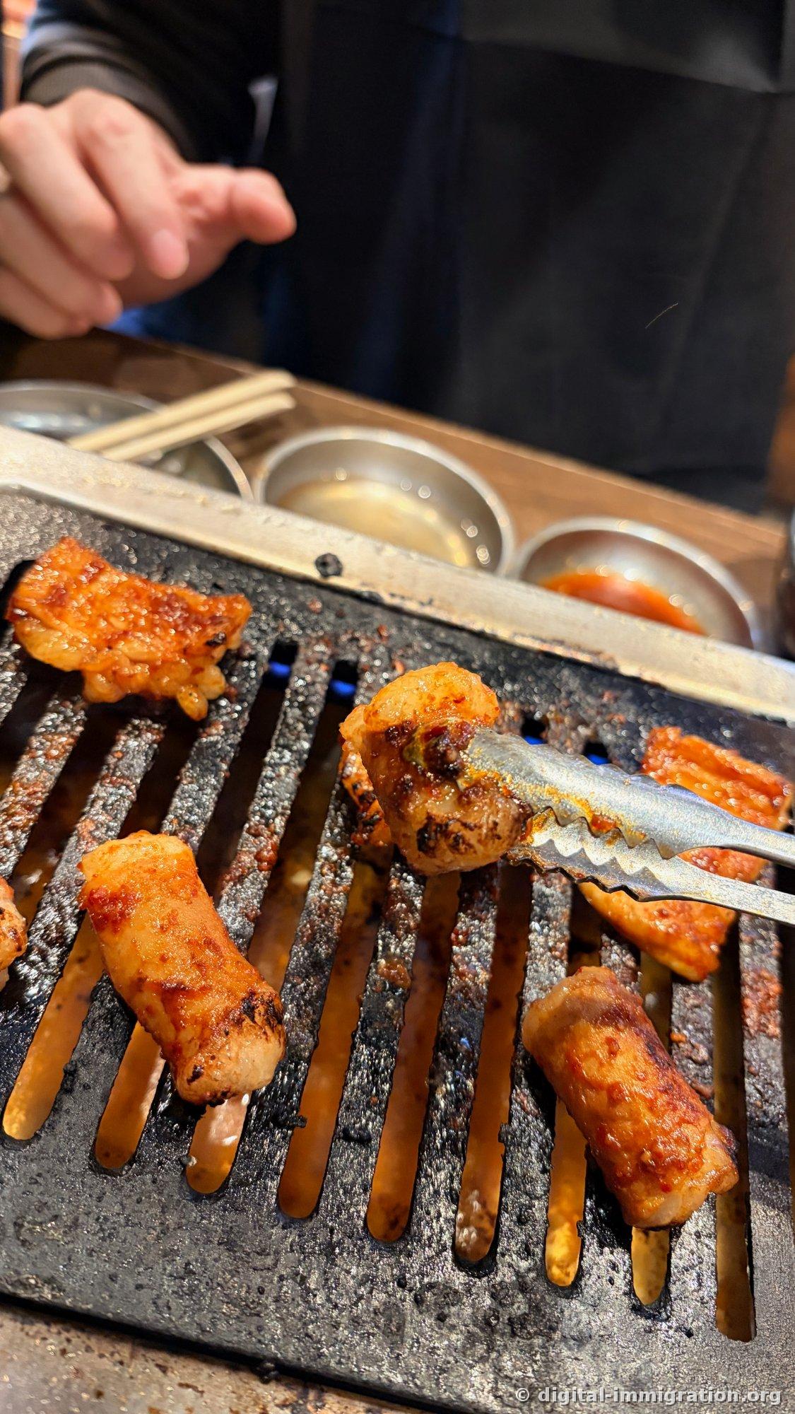 Seasoned hormon with a charred exterior being flipped with tongs; multiple pieces cooking on the grill with sauce dishes in the background