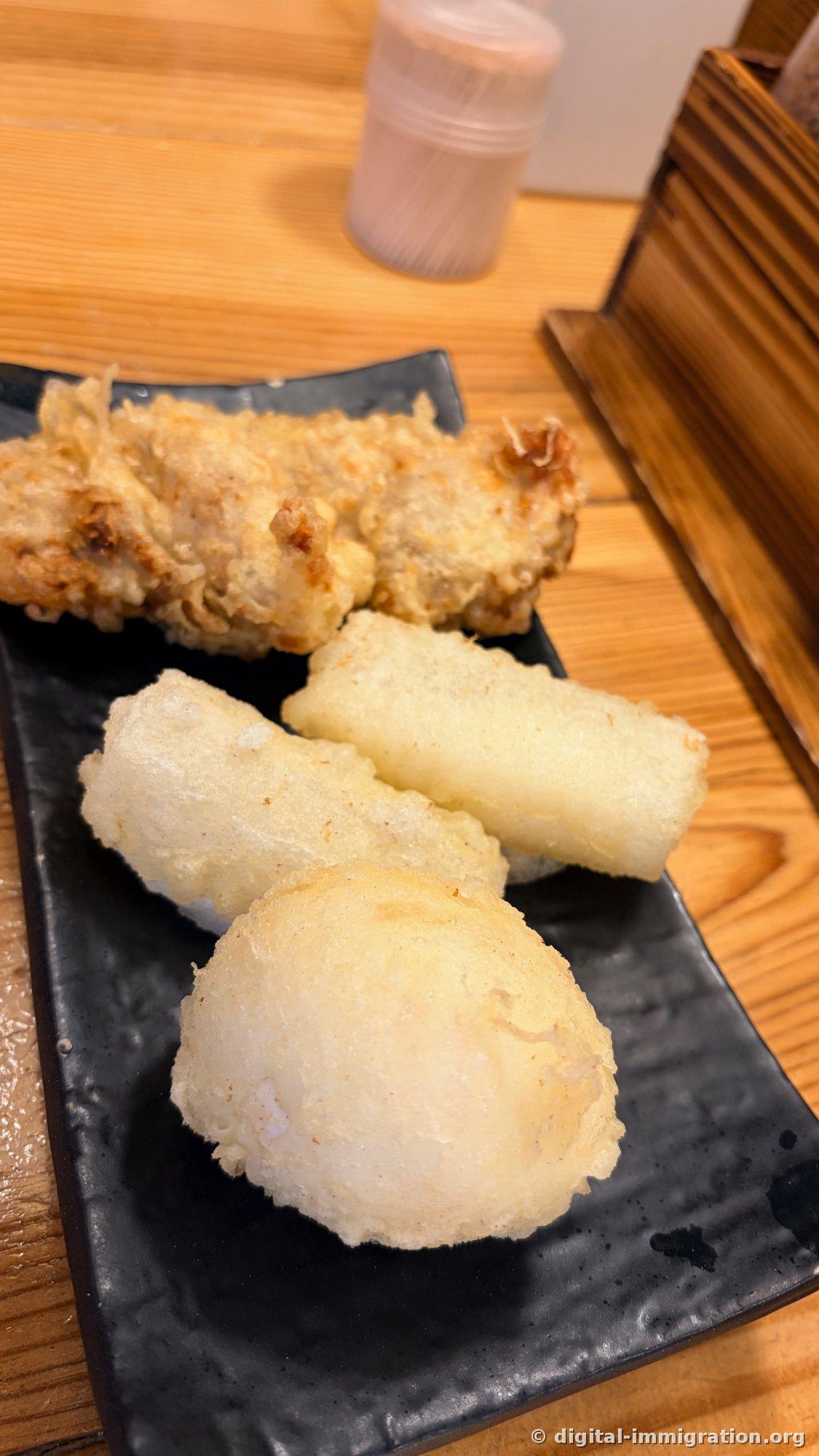 黒い四角い皿に盛られた天ぷら盛り合わせ。奥に鶏天ぷら、手前に白い揚げもち2個と丸い天ぷら1個が置かれている