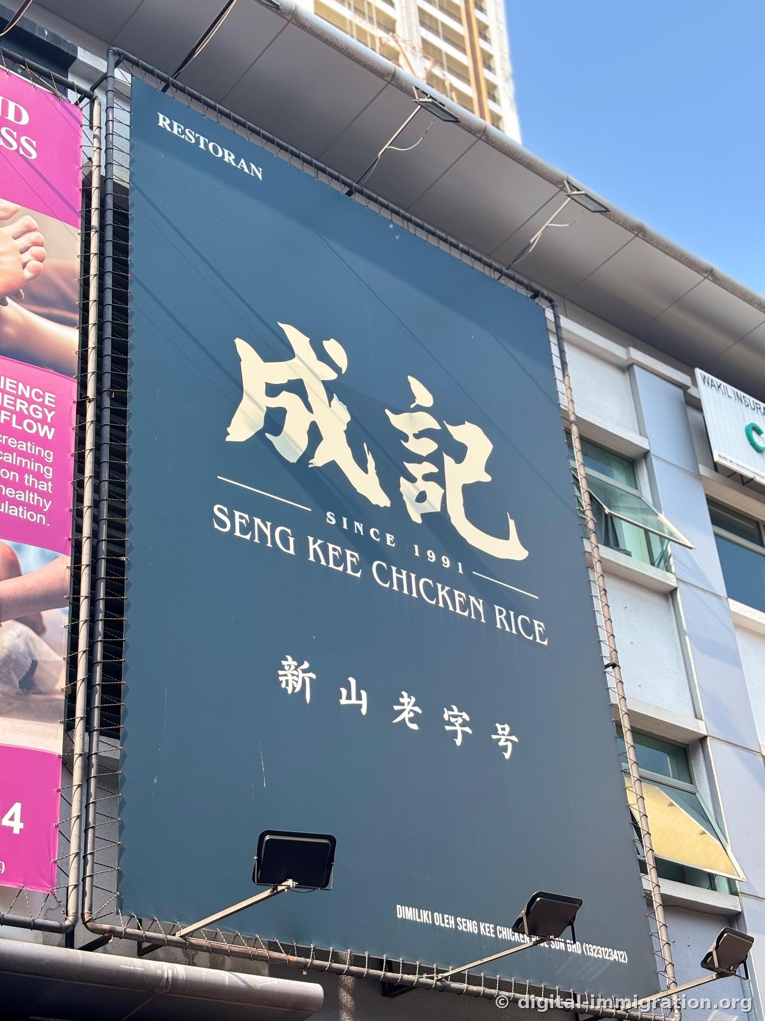 A large dark teal sign on the building exterior. Below large Chinese characters 成記 are SINCE 1991, SENG KEE CHICKEN RICE, and 新山老字号
