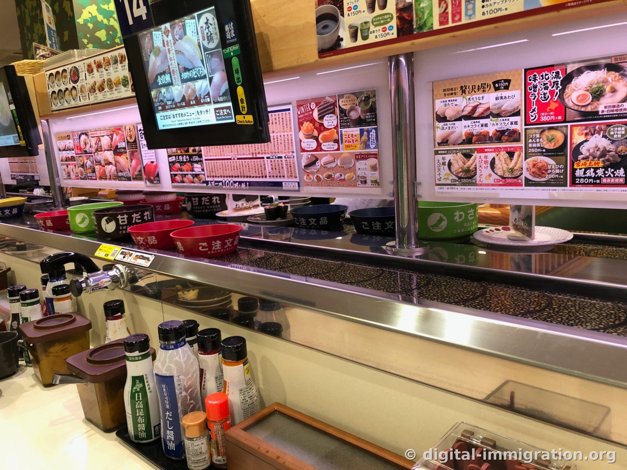 Au-dessus du convoyeur se trouvent un flacon de wasabi et un flacon de sauce sucrée, des assiettes de commande, et devant le comptoir se trouvent plusieurs bouteilles de sauce soja comme la sauce soja kombu Hidaka