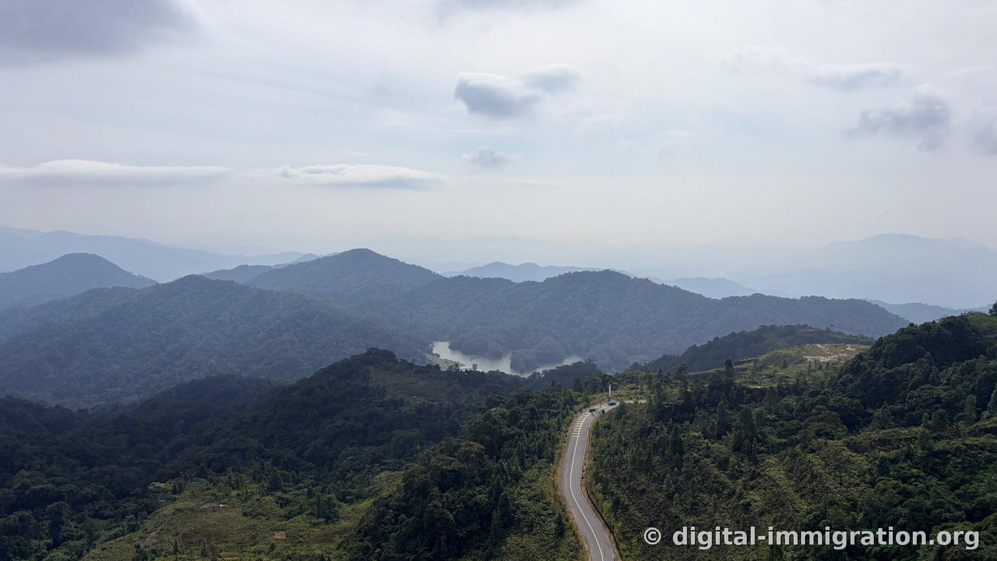 Wide panoramic mountain range with reservoir in center and mountain road continuing below