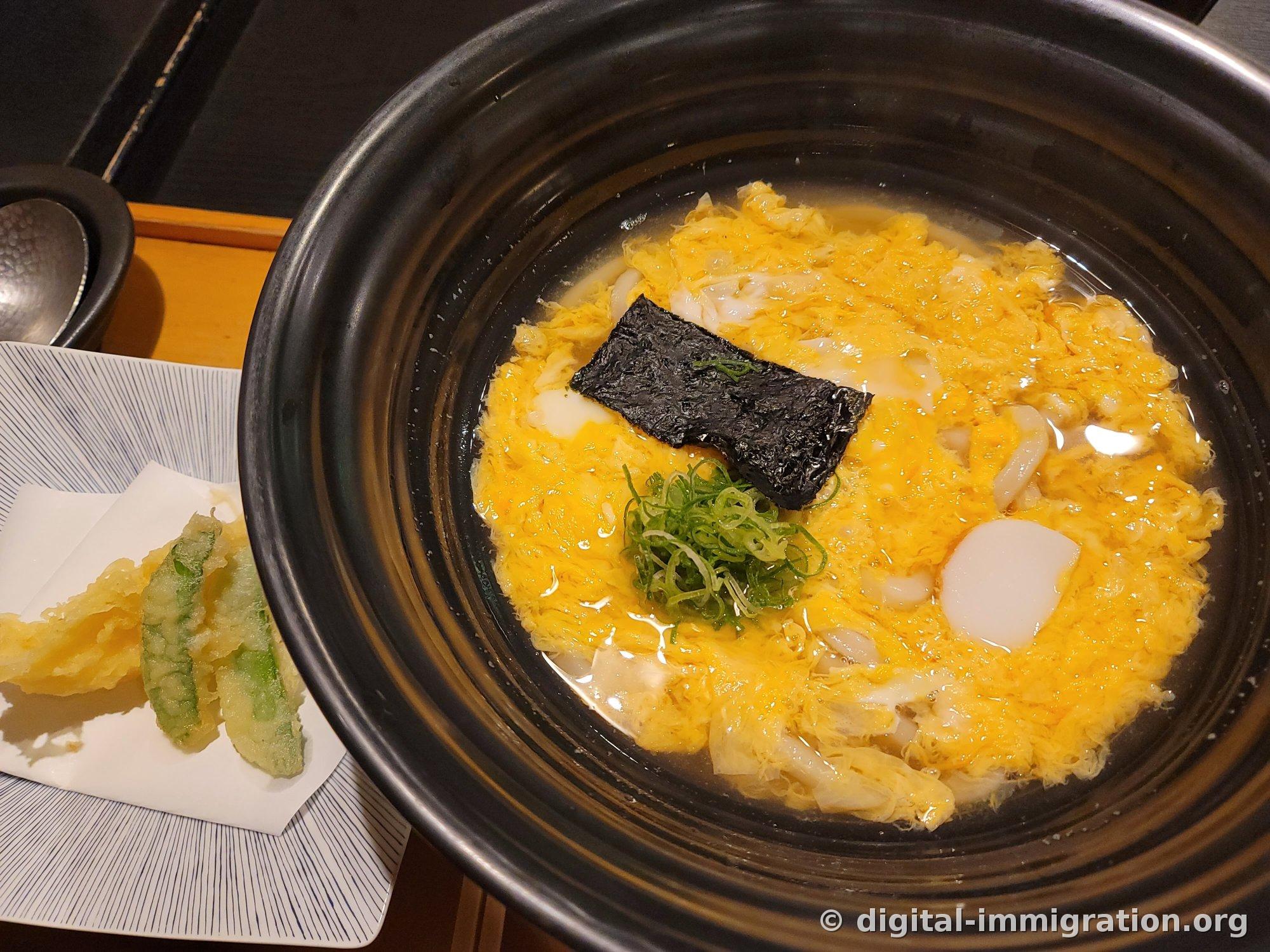 Un udon chaud aux œufs battus dans une cocotte noire. Les œufs jaunes, une feuille d'algue nori et de la ciboule sont garnis dessus, et un plat adjacent contient des piments shishito frits