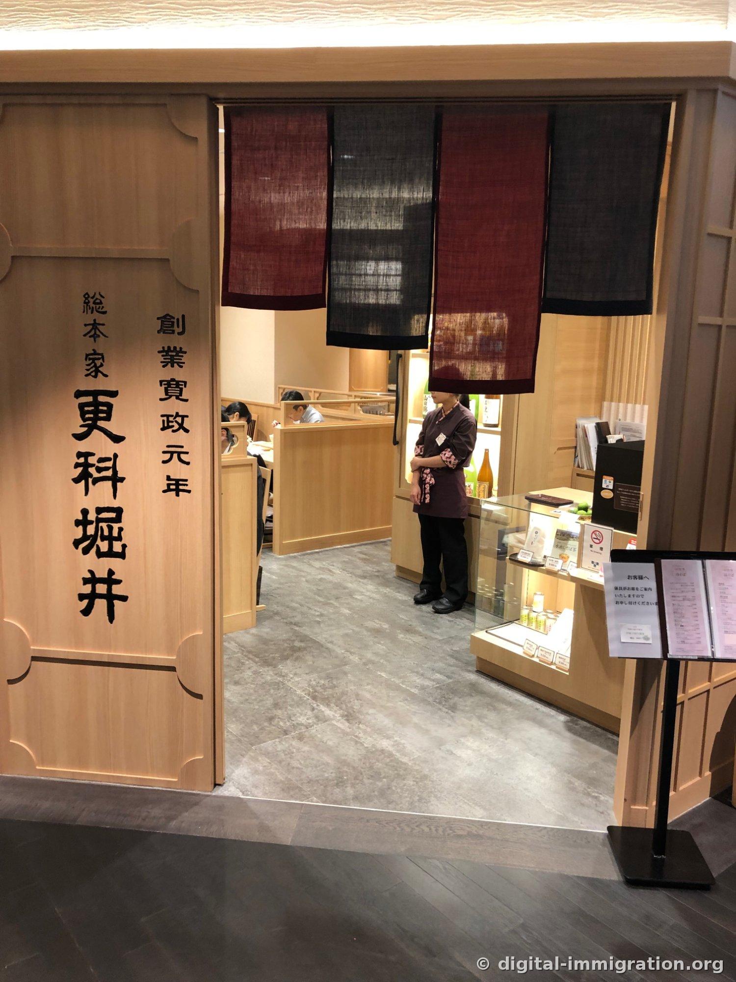 A signboard reading 'Sarashina Horii, Founded in Kansai Year One' in vertical script on a cypress wood wall, with maroon and black noren curtains at the shop entrance and a staff member standing