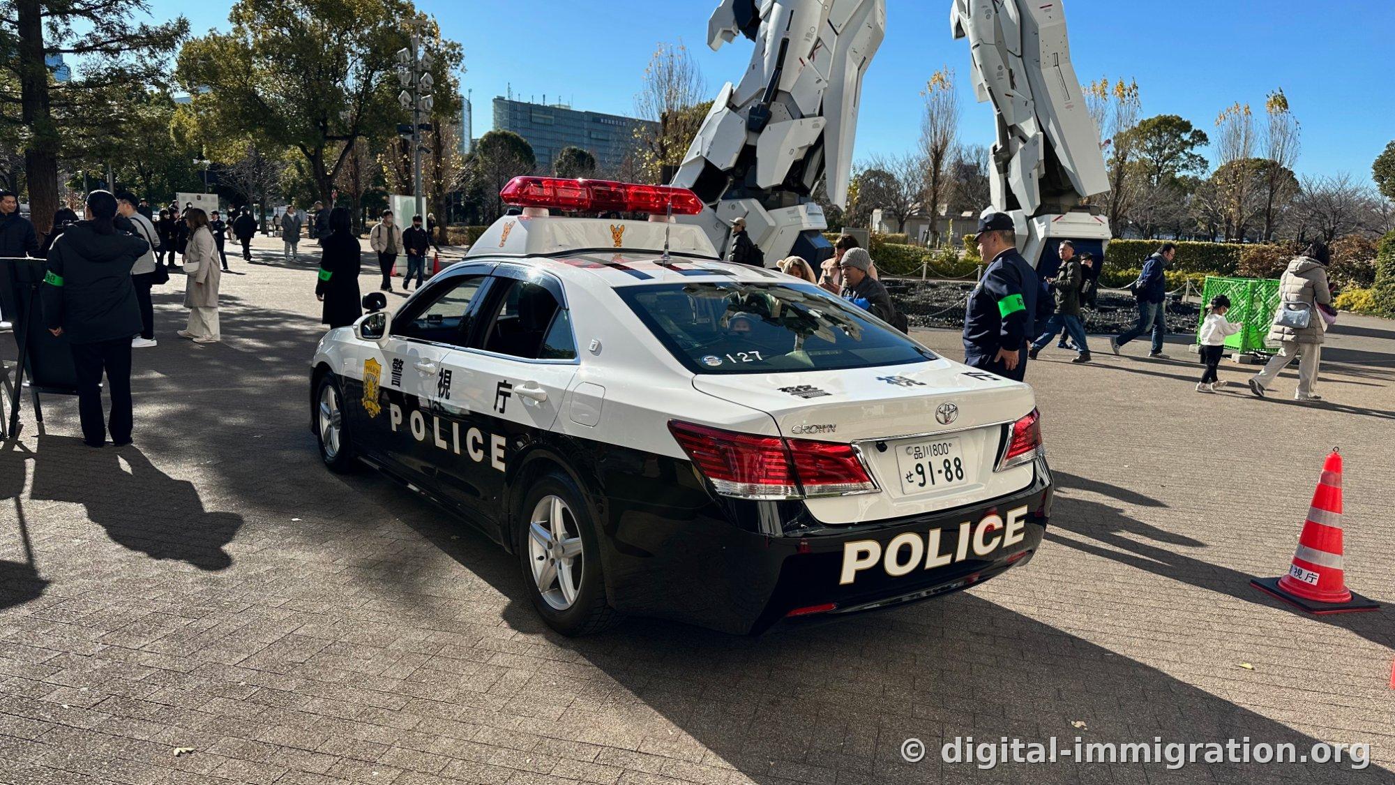 A black-and-white metropolitan police patrol car stationed in the plaza in front of the Gundam, tourists in winter clothing passing by, police officers with armbands and orange traffic cones