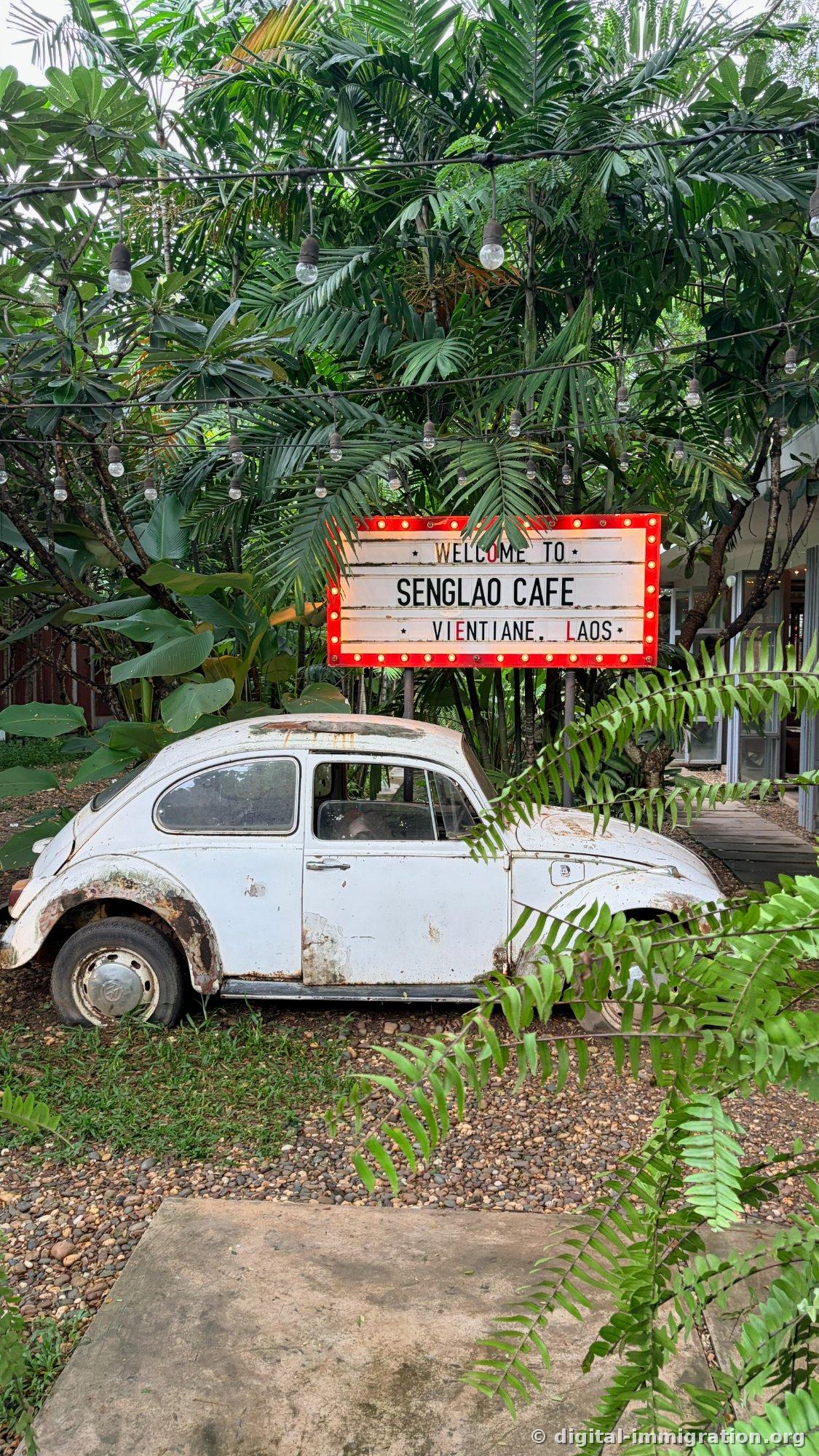 Une vieille Coccinelle Volkswagen blanche garée dans un jardin sous des cocotiers et des fleurs de plumeria, avec une guirlande lumineuse, une enseigne au néon au-dessus disant 'WELCOME TO SENGLAO CAFE VIENTIANE, LAOS'