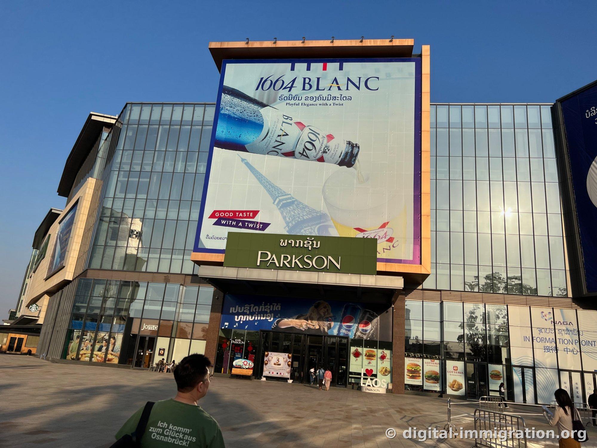 Vue générale de l'entrée du Parkson Mall sous un ciel bleu avec une grande publicité 1664 BLANC et l'enseigne PARKSON en caractères lao, des gens marchent sur la place devant