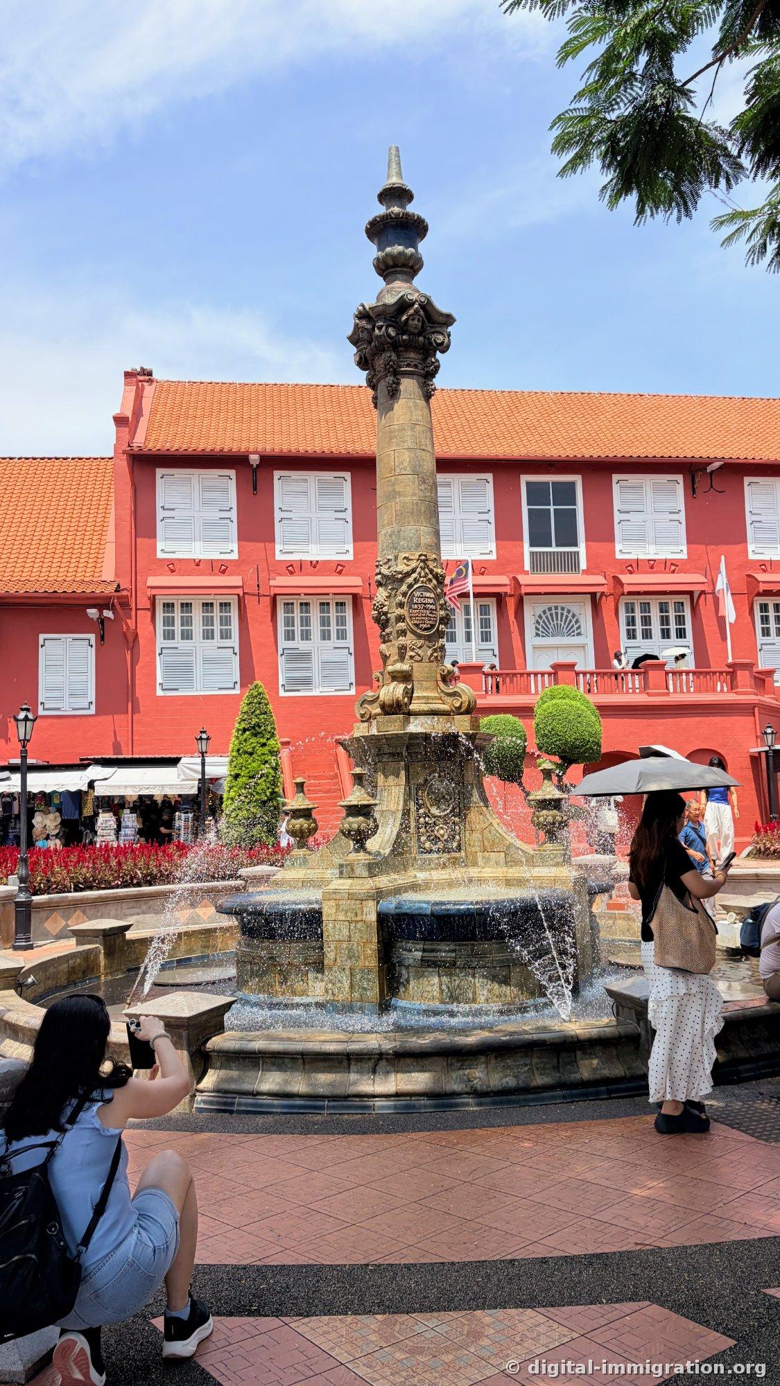 Victoria Fountain in front of the red Stadthuys building. A woman holding a black parasol stands on the right, and a tourist crouches to photograph the fountain on the left