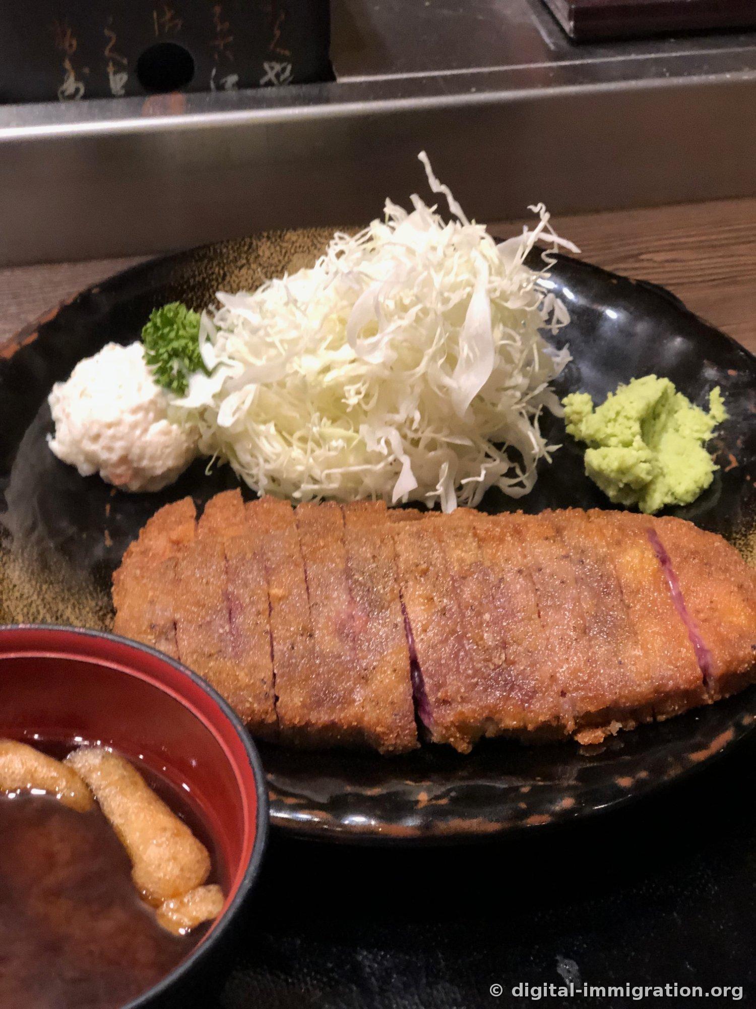 Un plateau complet de Gyukatsu teishoku dans une assiette noire. Côtelette de bœuf épaisses, chou émincé abondant, salade de pommes de terre et persil, wasabi, à côté un bol rouge avec miso soup contenant du tofu frit