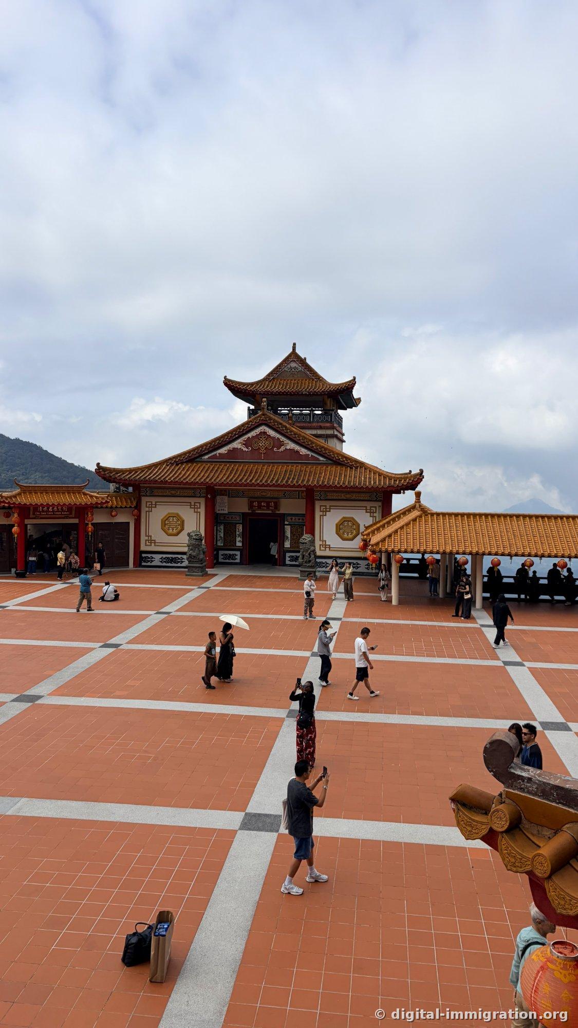 Vue de dessus d'une large place en carrelage orange et du bâtiment principal du temple à étage, une paire de lions en pierre devant, des touristes prenant des photos