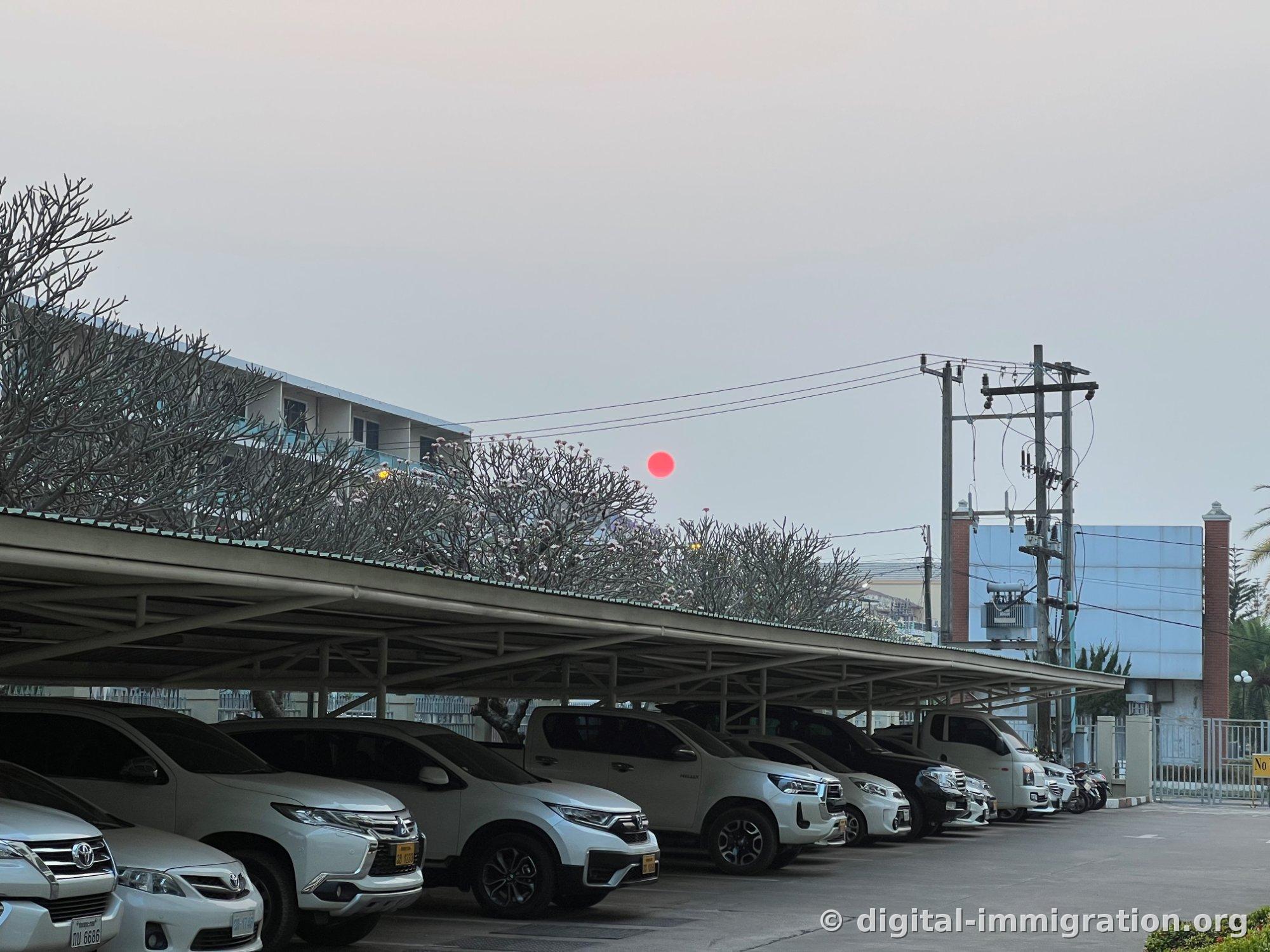 屋根付き駐車場に白いSUVとピックアップトラックが並べられており、プルメリアの木越しに赤い夕日が沈む夕方の風景