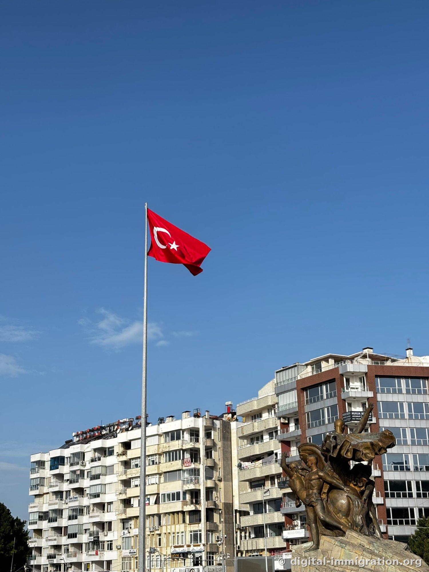 A Turkish flag hanging high against a blue sky with the back of the bronze equestrian statue visible below and to the right, high-rise apartments with air conditioning units and shop signs in the background.