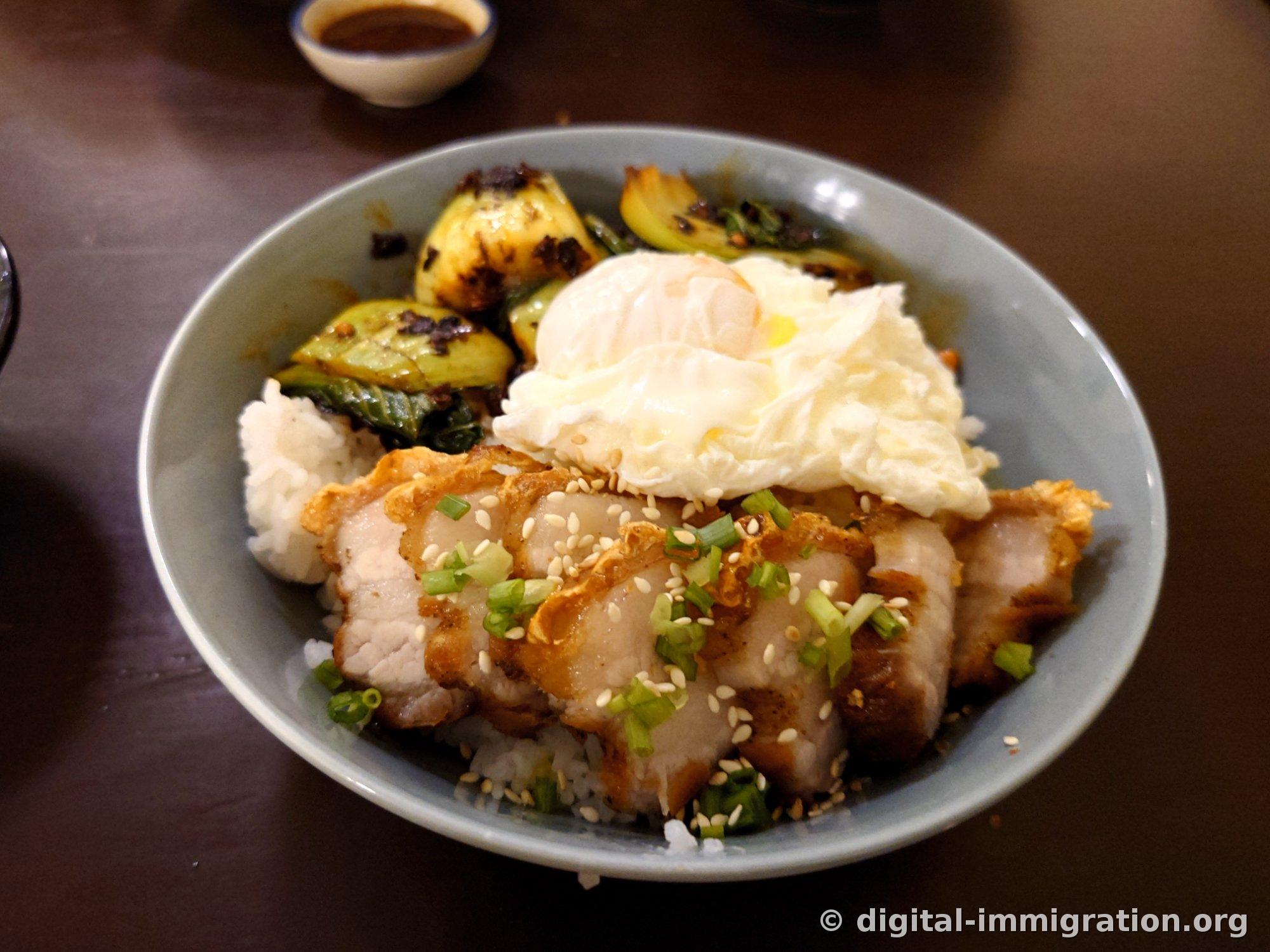 白いご飯の上に皮がカリッとした豚バラ肉のスライスと温泉卵、炒めた青菜を乗せた丼。ゴマと細ネギが振りかけられている