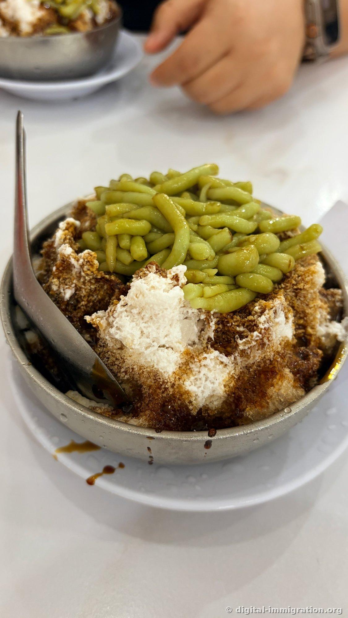 One bowl of cendol in a stainless steel bowl. Finely shaved ice is topped with thick green pandan jelly noodles with brown palm sugar syrup dripping down, with a spoon inserted. A second identical bowl is visible on the white table behind.