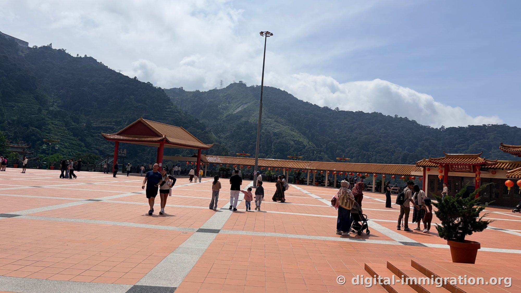 Un pavillon marqué 'HUA HALL' et un long corridor avec des lanternes rouges suspendues, une crête montagneuse enveloppée de nuages en arrière-plan