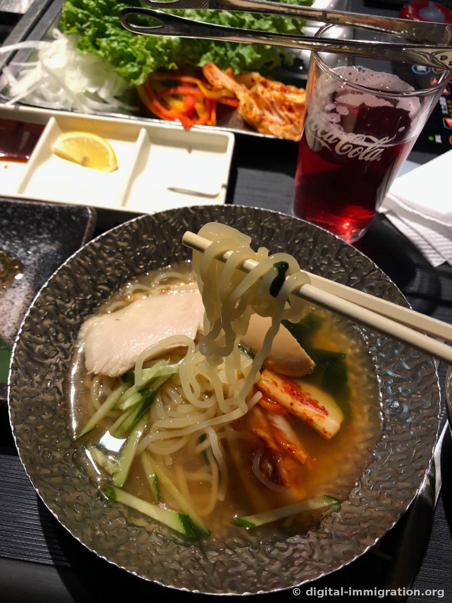 银色碗里的冷面被筷子夹起，上面放着叉烧片、切丝黄瓜和泡菜，后面看得到可乐杯和生菜盘子