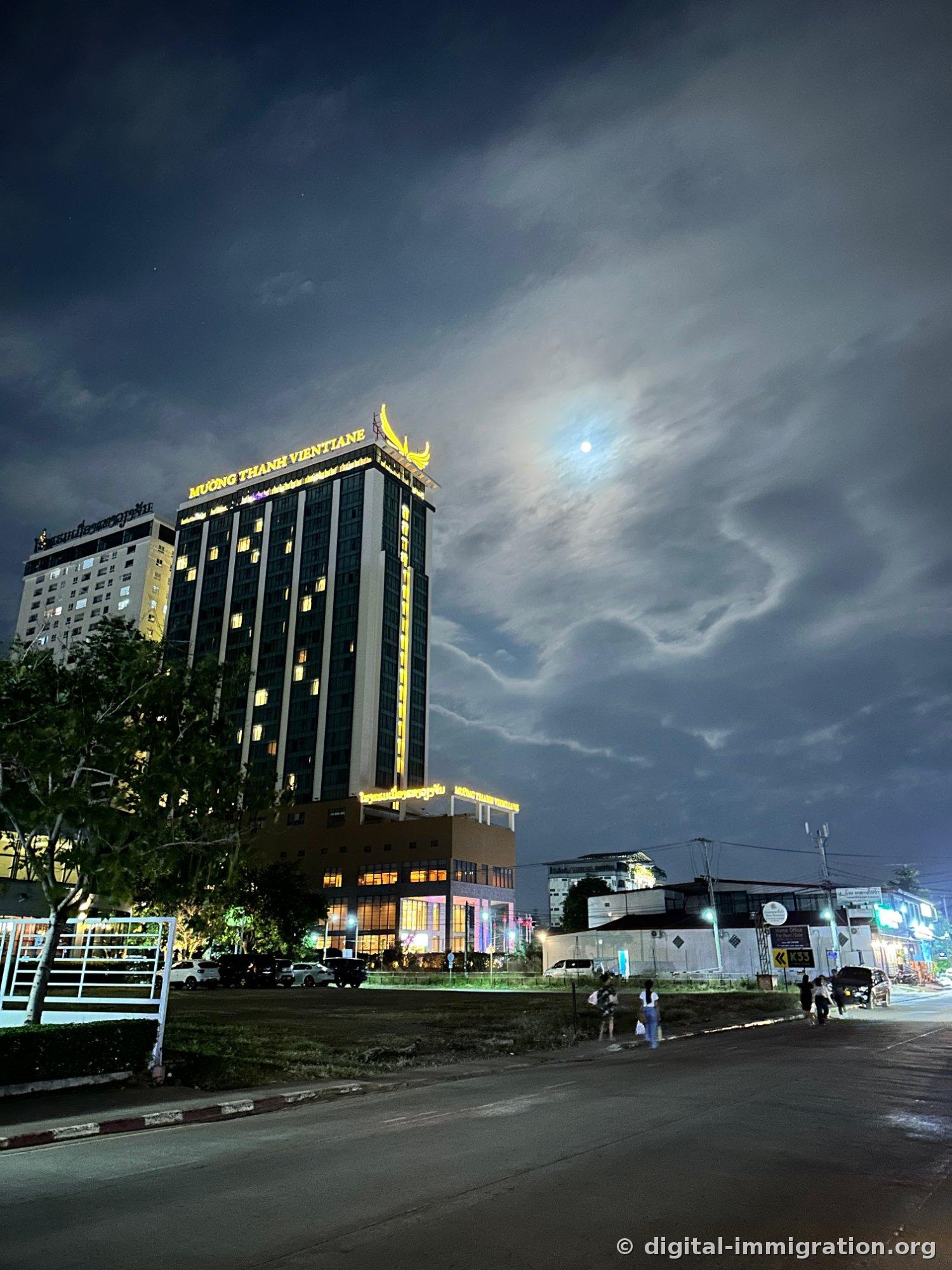 Vue nocturne du Mường Thanh Vientiane depuis l'autre côté de la route, avec l'enseigne jaune « MUONG THANH VIENTIANE » allumée sur le toit et les ailes dorées de l'oiseau