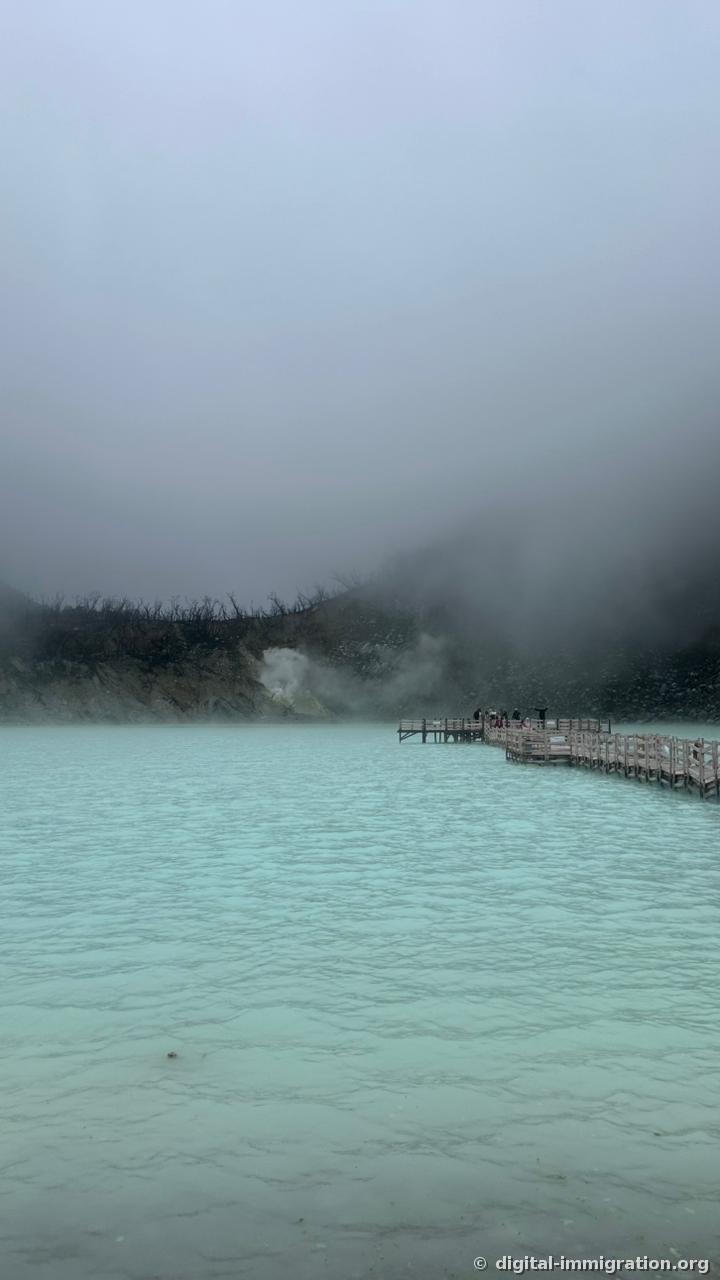 A vertical photo taken close to the water surface, with the wooden deck continuing over the cyan water and white sulfur steam rising from rocks on the far side