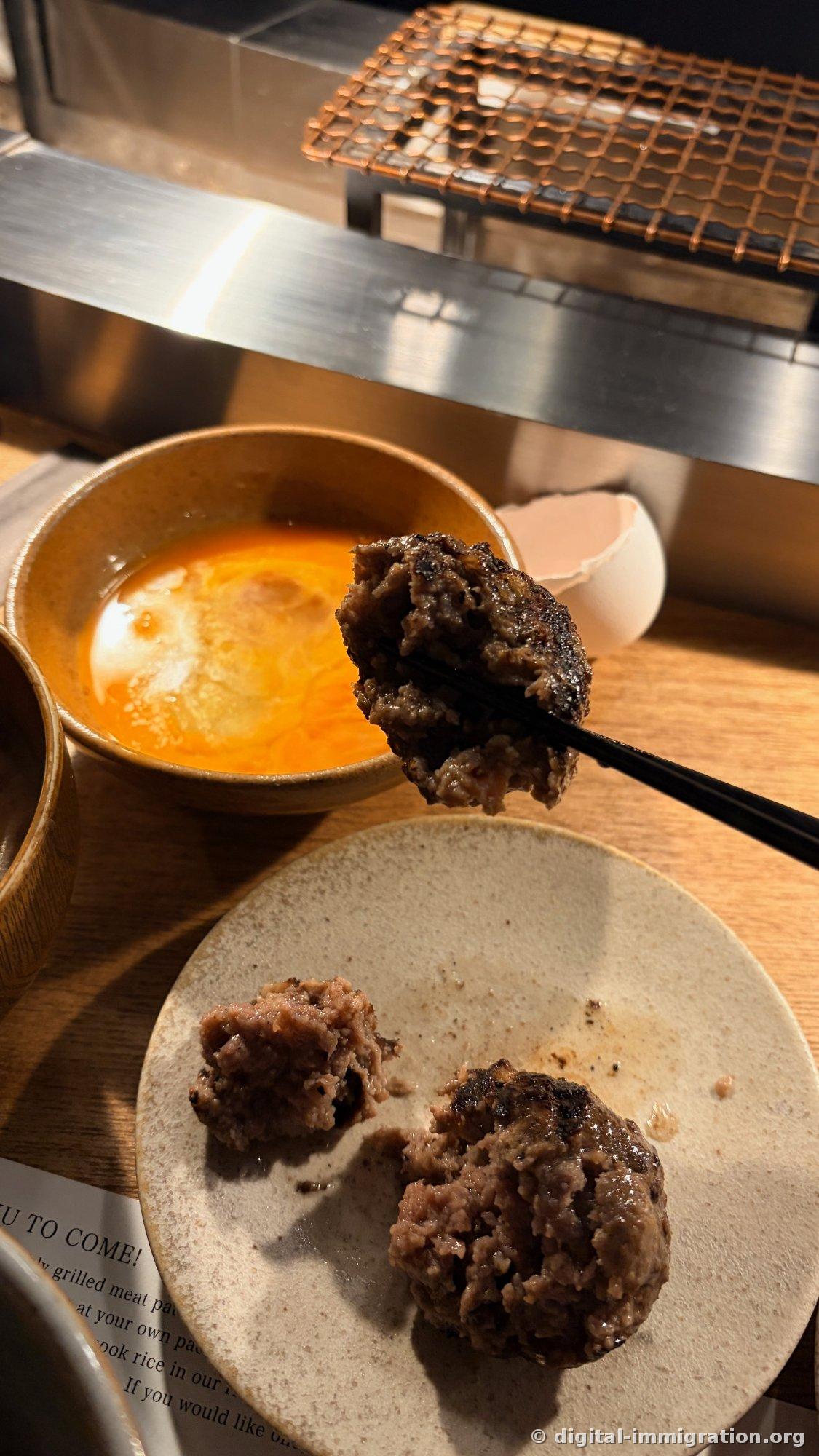 Des baguettes tenant un hamburger juste avant de le tremper dans le bol de sauce à l'œuf, et dans l'assiette devant, un hamburger coupé en deux montrant l'intérieur
