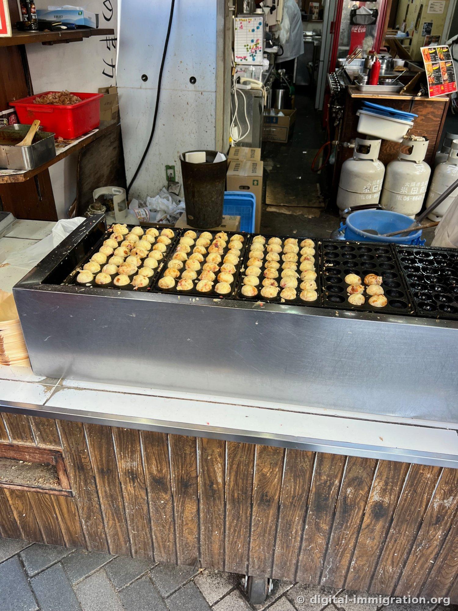Douzaines de takoyaki qui dorent sur une longue plaque de fer d'un petit restaurant, avec la cuisine et les bidons de gaz derrière