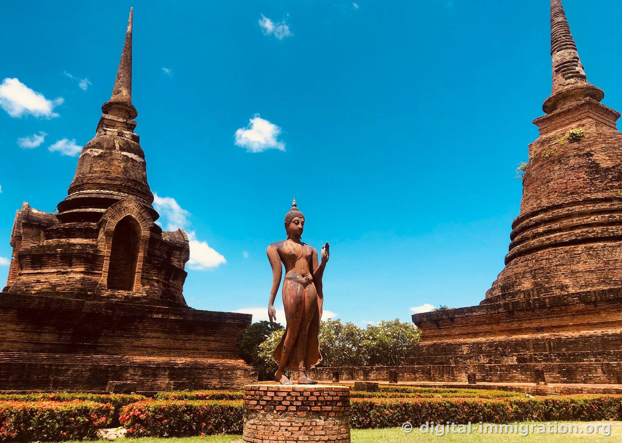 A walking Buddha statue standing on a circular platform between two brick spires, with right hand raised and a red flower bed at its feet
