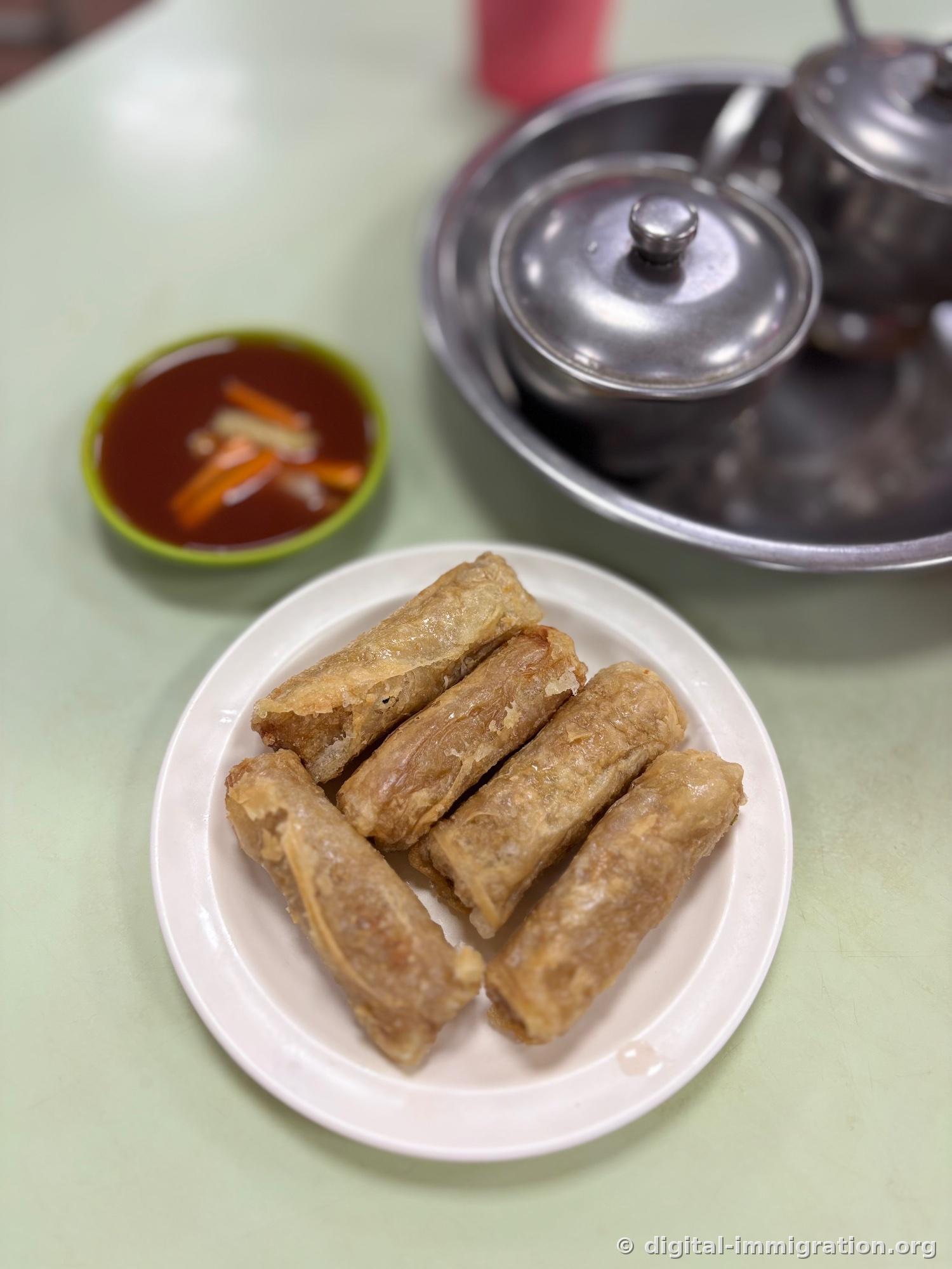 Assiette blanche contenant cinq rouleaux de tahu kulit frits avec coupelle de sauce rouge garnie de carotte, pot de sauce couvert sur plateau inoxydable en arrière-plan