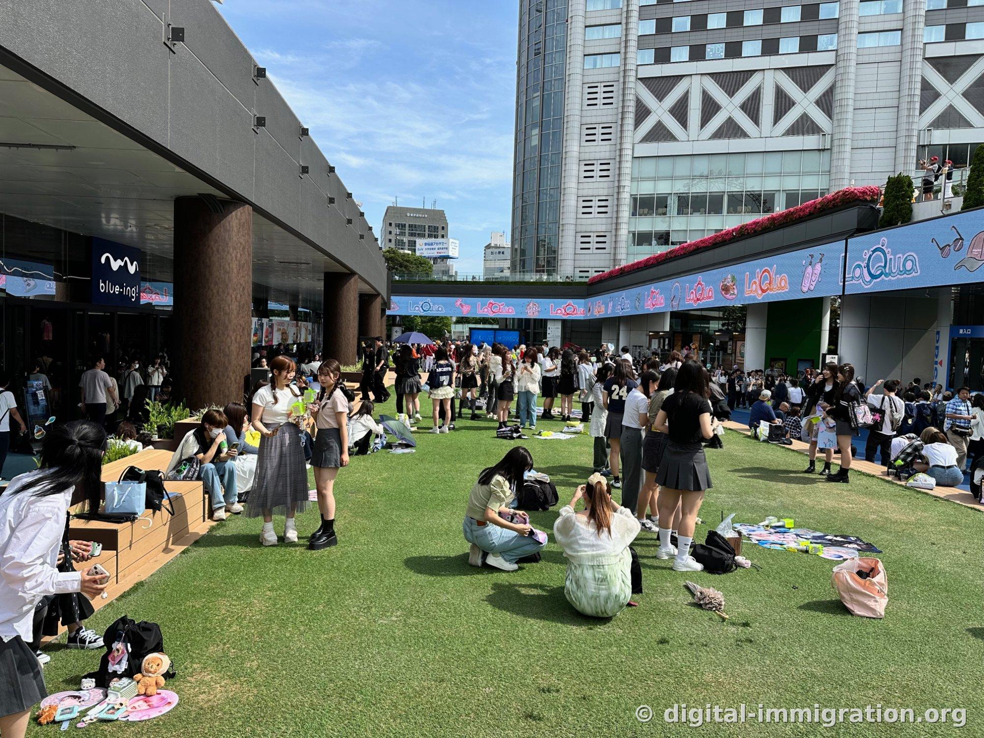 Devant le bâtiment avec l'enseigne bleue LaQua, de jeunes gens sont assis ou debout en train de prendre des photos sur la pelouse artificielle, et on voit un tapis rose avec des figurines et des articles de collection posés sur l'herbe à l'avant