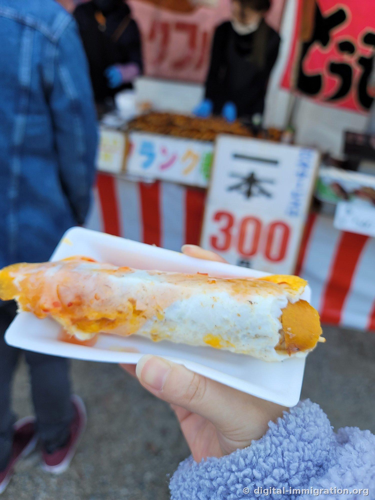 Une main tenant un hot-dog enrobé d'œuf sur une assiette en papier, avec un panneau de vendeur ambulant indiquant 300 yens pour un en arrière-plan