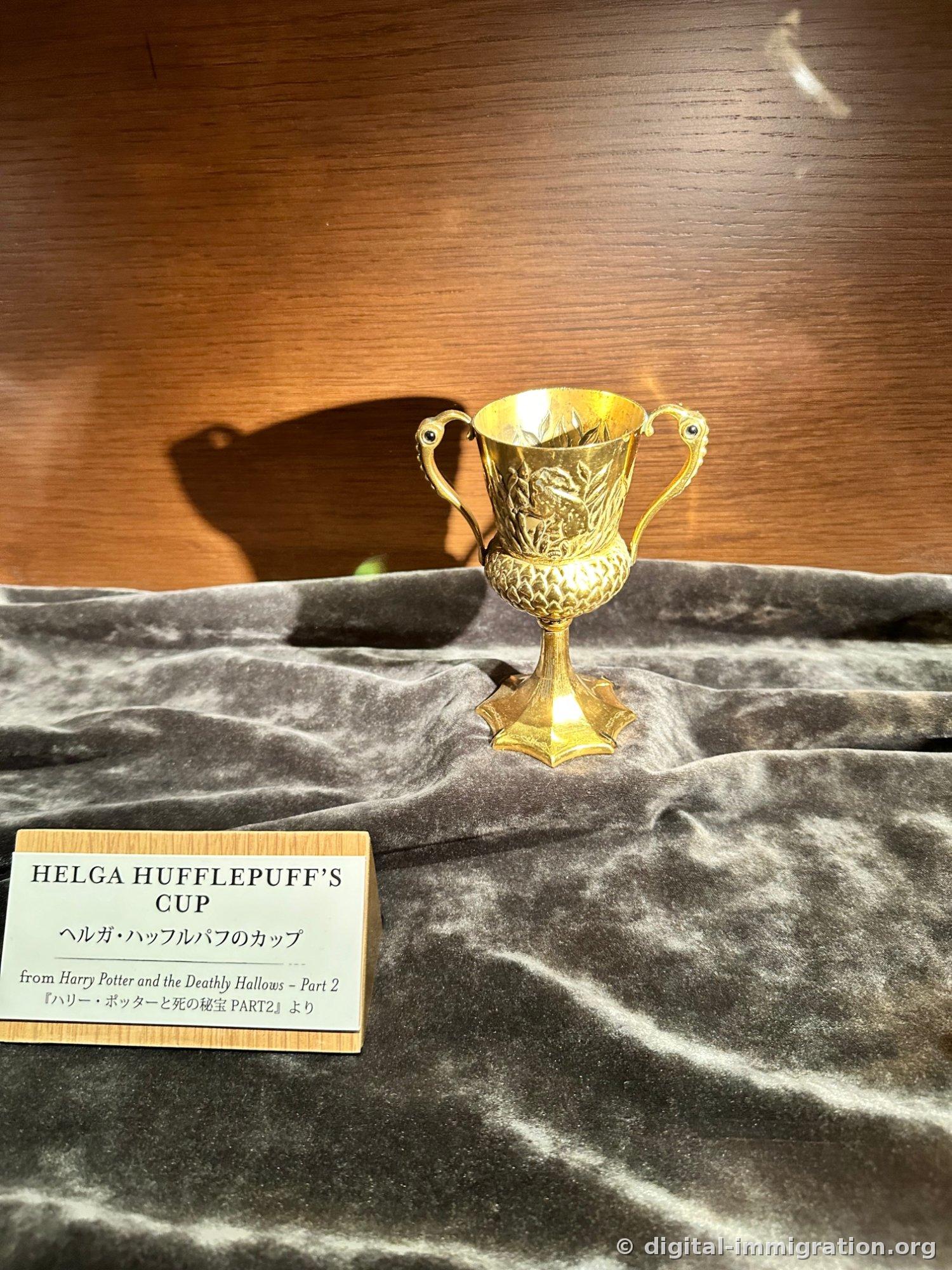 A small gold cup on gray velvet with a large shadow cast on the wooden wall behind, with the nameplate HELGA HUFFLEPUFF'S CUP