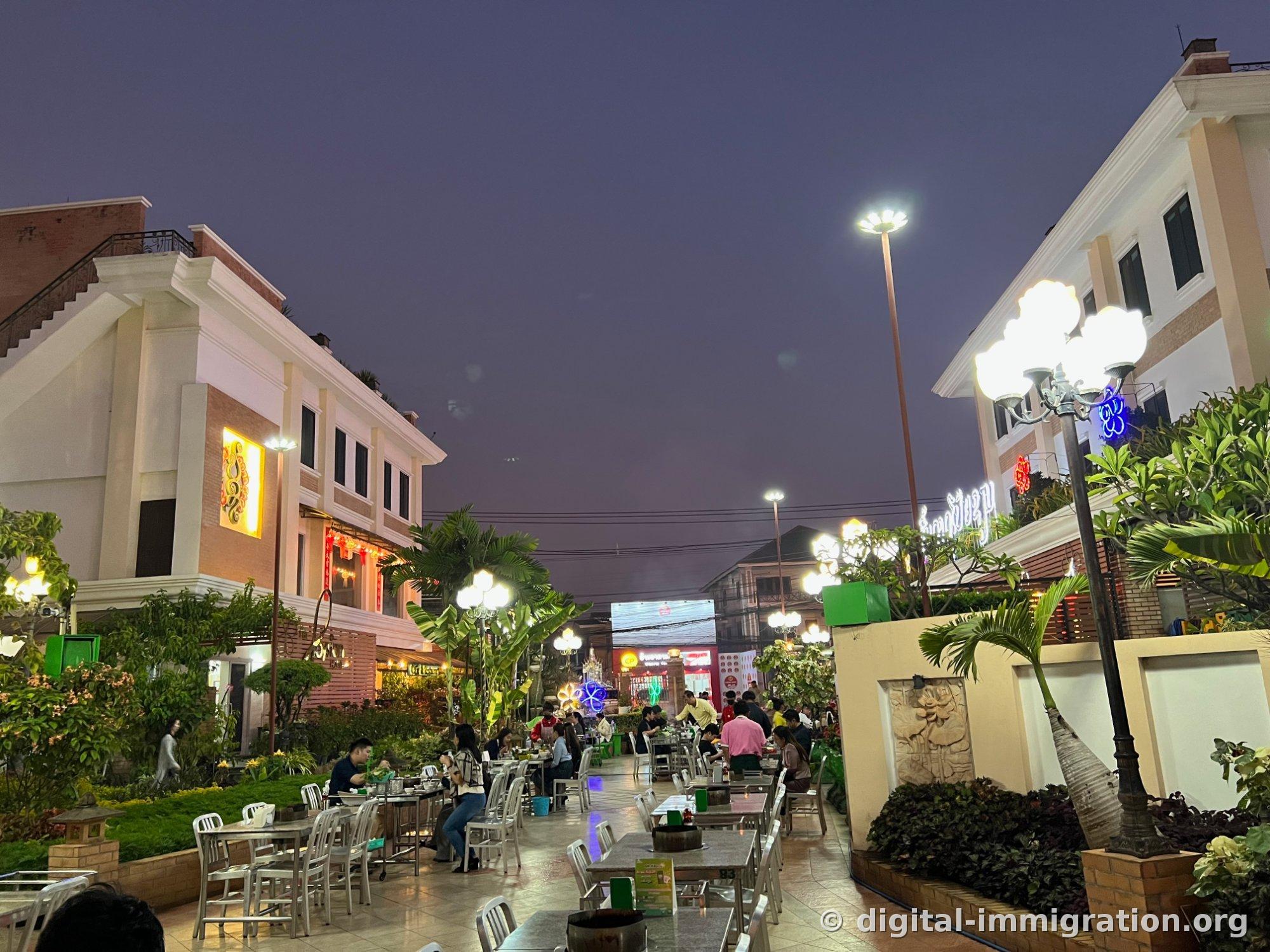An outdoor courtyard at night with stainless steel tables and white chairs in a row, and local families dining at Poysian