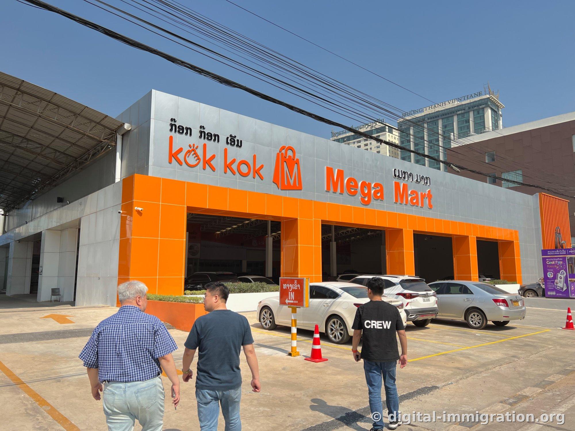 파란 하늘 아래 은색 외벽에 주황색 'kok kok Mega Mart' 간판이 붙은 건물 정면과 앞쪽 주차장, 뒤로 므앙탄 비엔티안 호텔이 보이는 전경