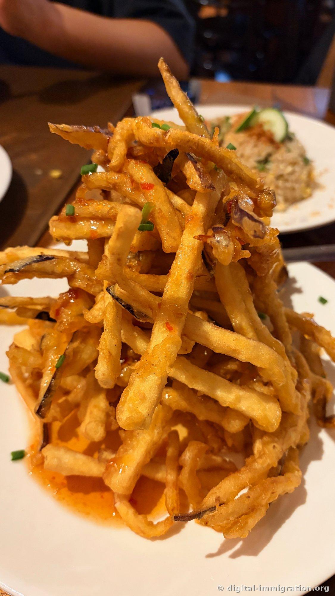 A pile of julienned eggplant tossed in red chili sauce, stacked high on a plate and topped with scallions, with a fried rice dish visible in the background