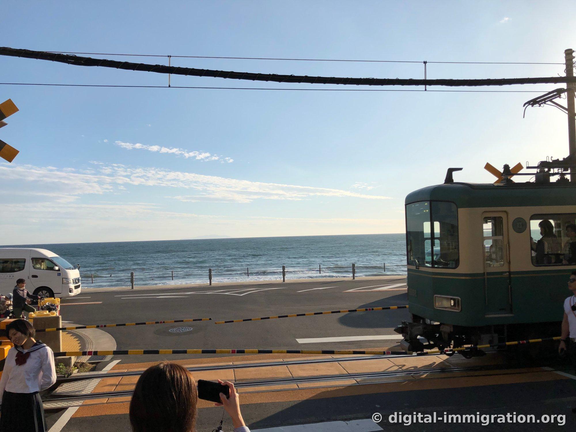 L'avant du tramway Enoden vert et crème entre dans le passage à niveau par la droite, avec une personne en costume marin à gauche