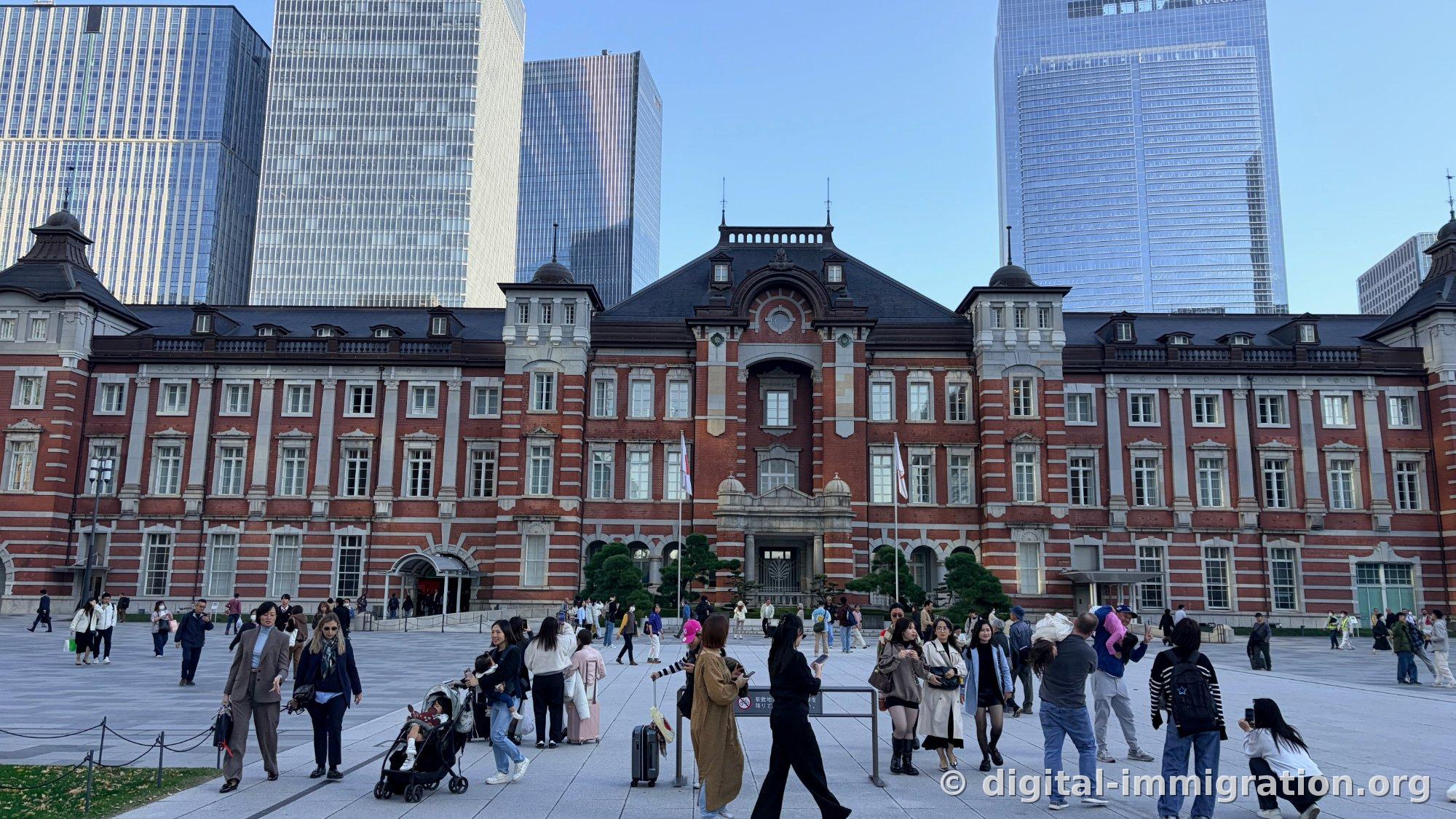 Vue de face complète du bâtiment en brique rouge de la Gare de Tokyo Marunouchi, avec deux drapeaux devant l'entrée principale et la place remplie de monde