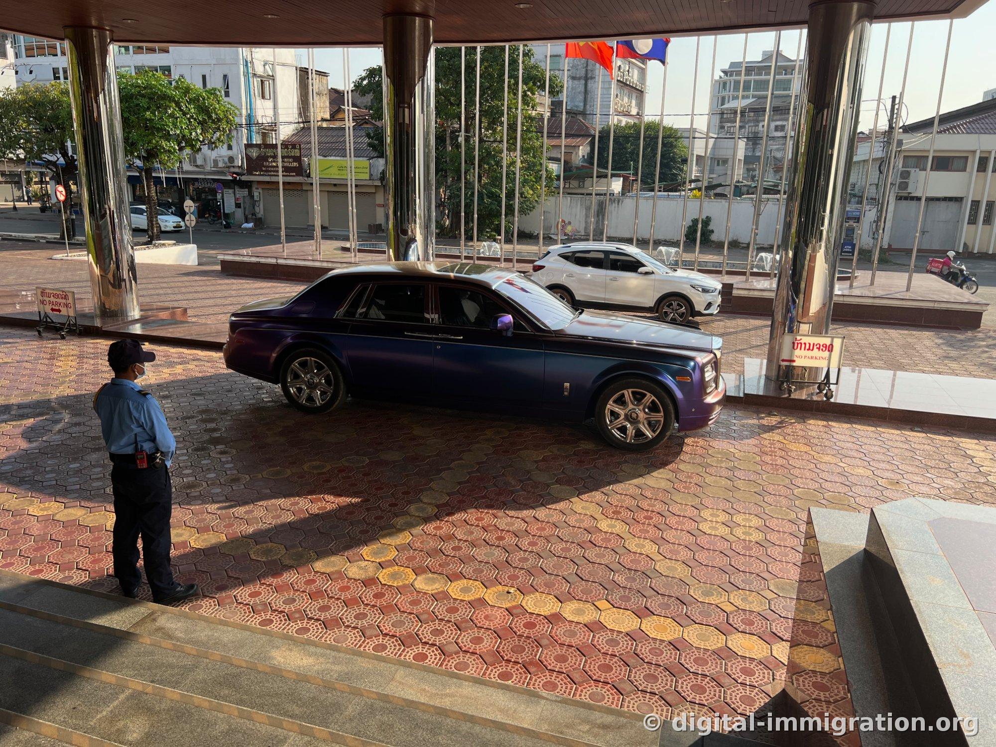 Rolls-Royce Phantom violet garé sous l'auvent de l'entrée principale avec un agent de sécurité en uniforme et drapeaux laotiens en arrière-plan