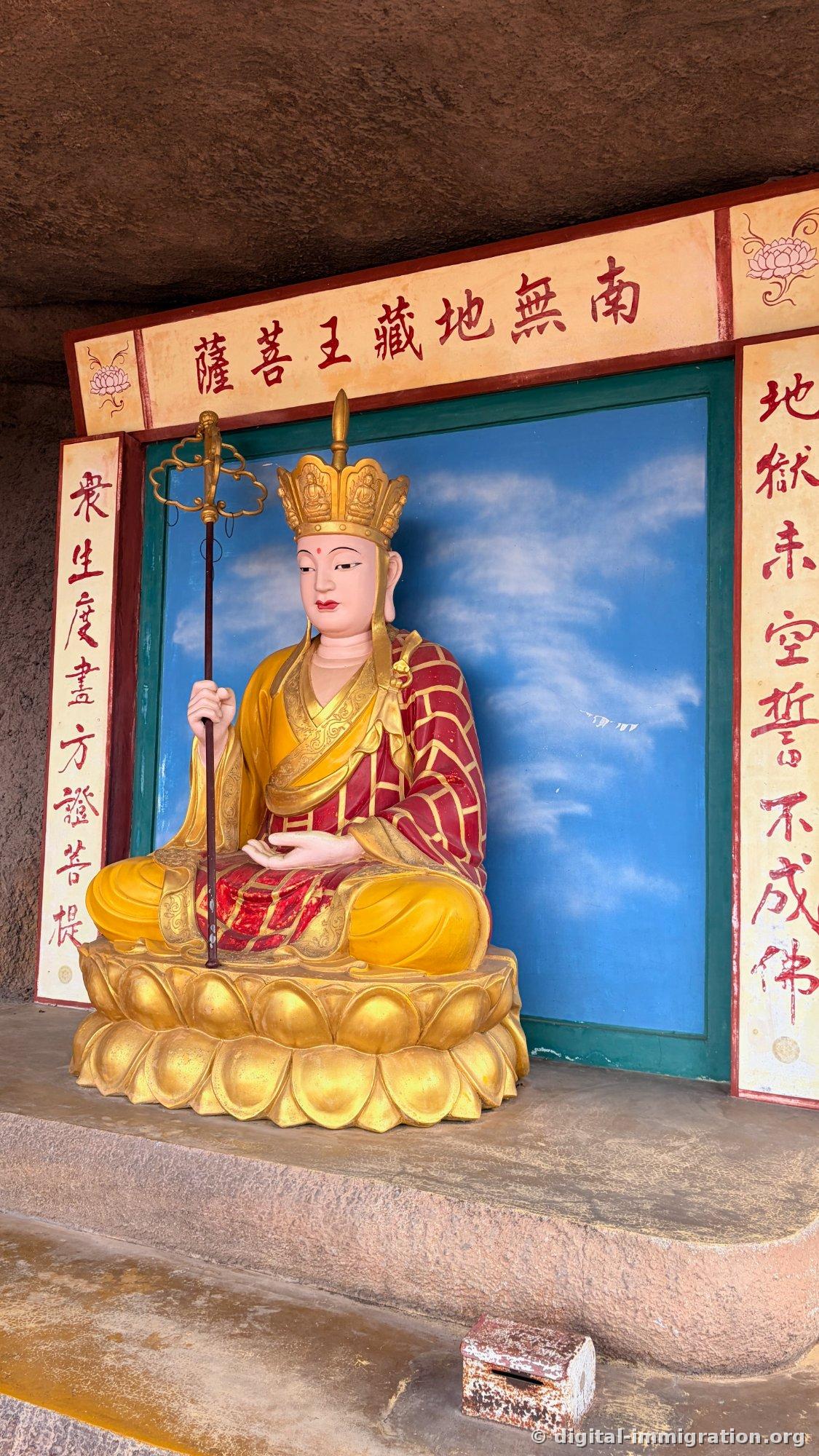 Statue asise dorée du Bodhisattva Ksitigarbha, tenant un bâton de moine, abritée dans un renfoncement rocheux avec des voeux écrits en caractères rouges sur les colonnes de chaque côté