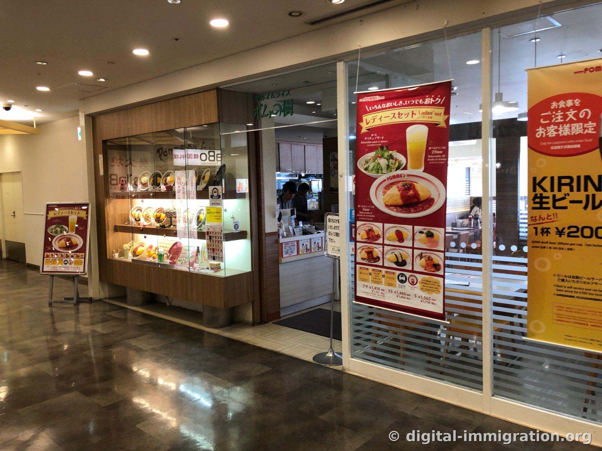 L'entrée du Pomme no Ki succursale Odaïba dans le couloir du centre commercial. On voit l'enseigne '創作オムライス ポムの樹' en lettres vertes, une banderole de promotion pour les femmes en rouge, et une banderole jaune de KIRIN Bière pression ¥200