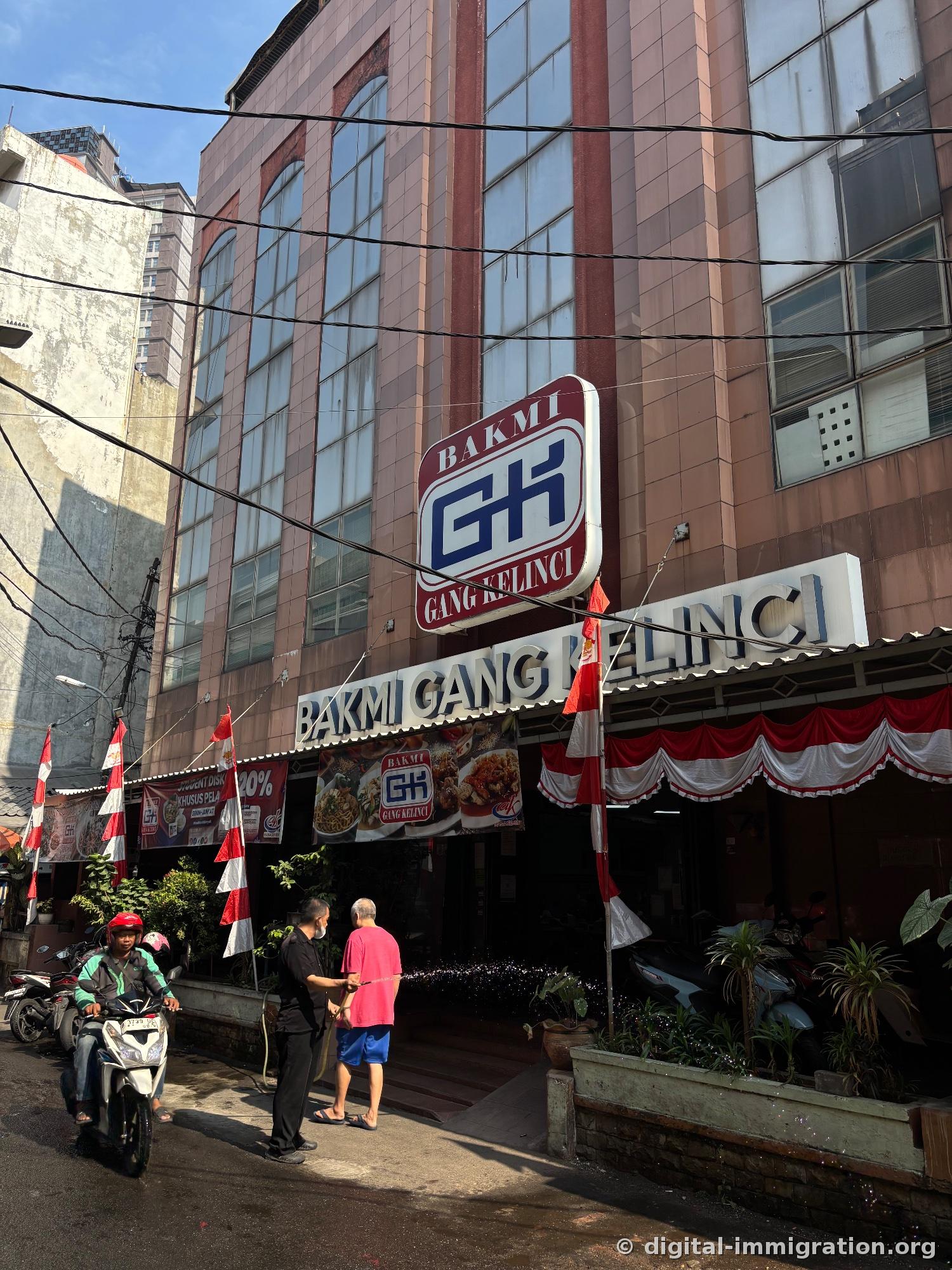 Devanture de 'BAKMI GANG KELINCI' sous le soleil du matin avec les drapeaux indonésiens rouges et blancs, des gens qui nettoient à l'eau et des motos garées devant