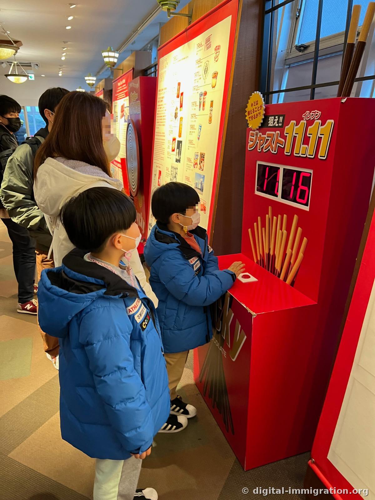 Children in front of a red game machine decorated with Pocky shapes, attempting to hit 11.11, with 11.16 displayed on screen