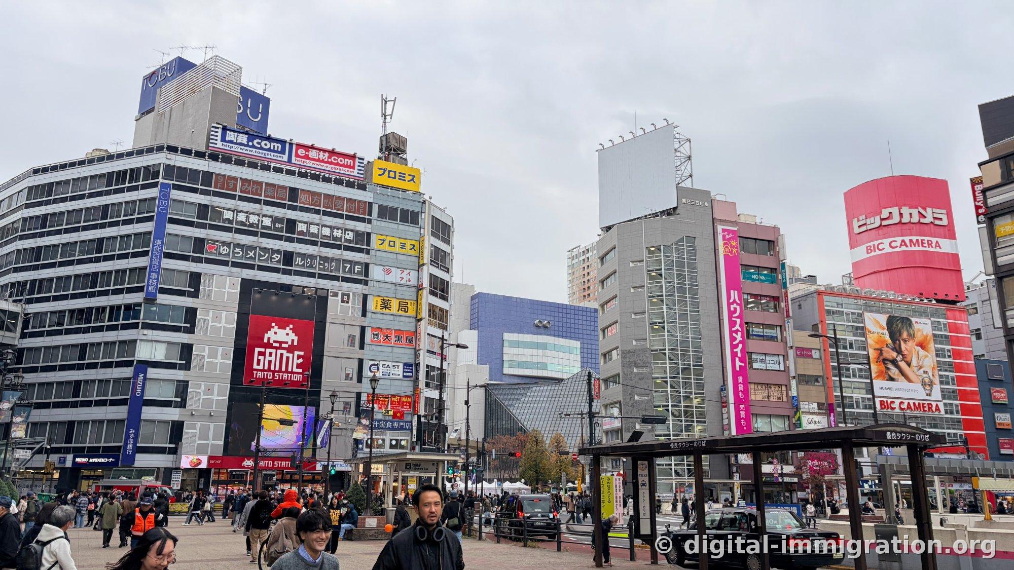 Ikebukuro Station plaza under an overcast sky; Tobu Department Store and Taito Station arcade signs visible, with Bic Camera's red sign and taxi stand on the right