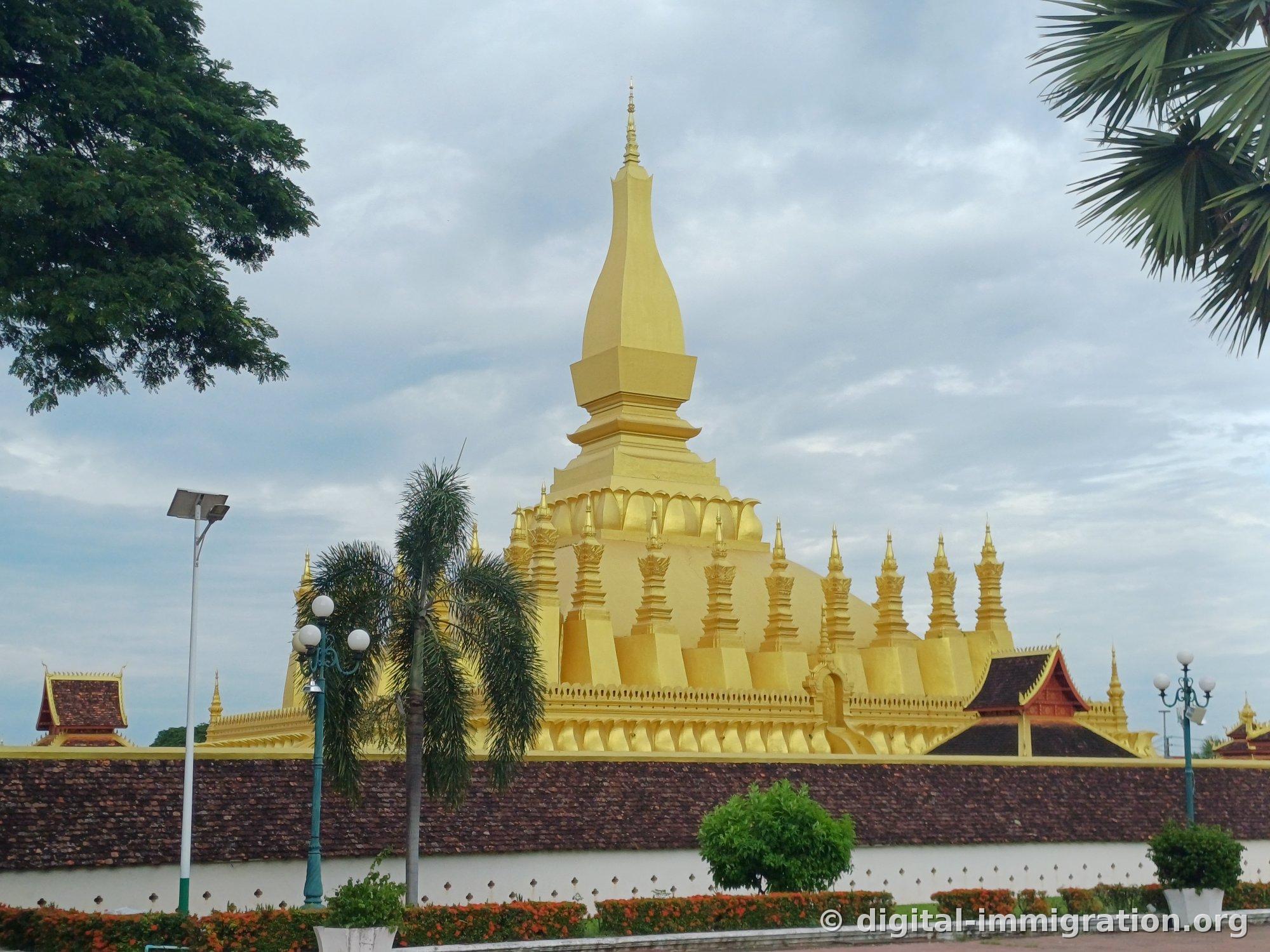Pha That Luang's golden stupa and palm trees rising beyond the wall under a cloudy sky