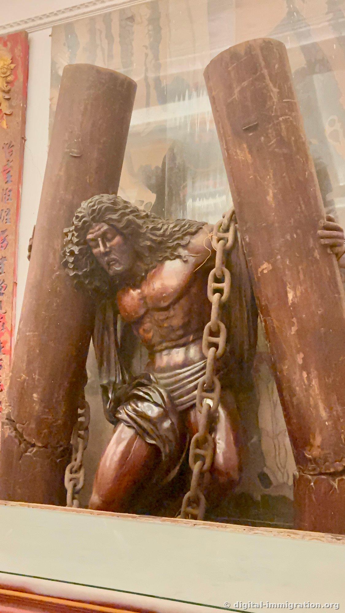 European-style wooden sculpture of a man on his knees bound with chains between two pillars inside glass display case