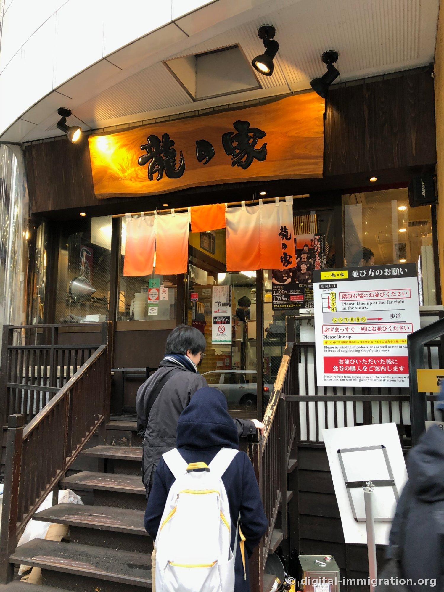 L'entrée de la succursale Shinjuku de Tatsunooya avec une enseigne gravée 龍の家 sur une planche de bois et un noren orange, avec deux clients montant l'escalier