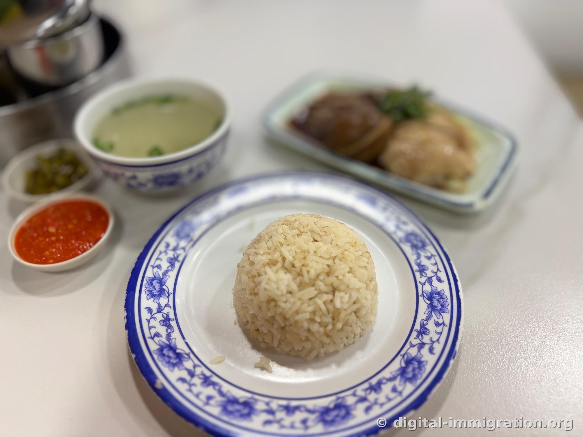 Blue and white porcelain plate with oil rice molded in a half-sphere, beside it red chili sauce and pickled green chilies, clear broth, and the chicken plate in back