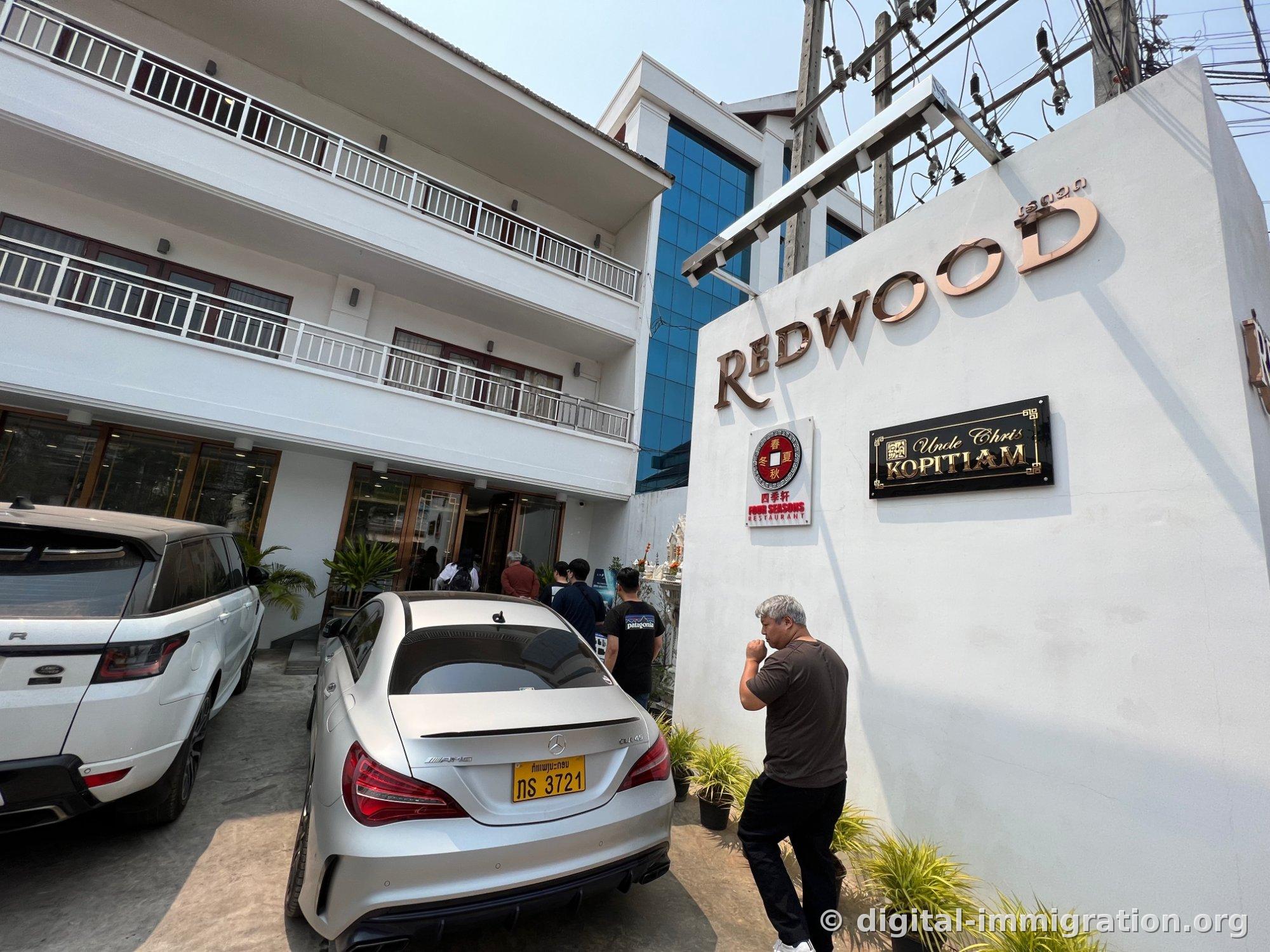 Restaurant entrance with a white brick wall displaying the rose gold REDWOOD sign, Four Seasons Restaurant and Uncle Chris Kopitiam nameplates, and a white Mercedes and Range Rover parked in the courtyard