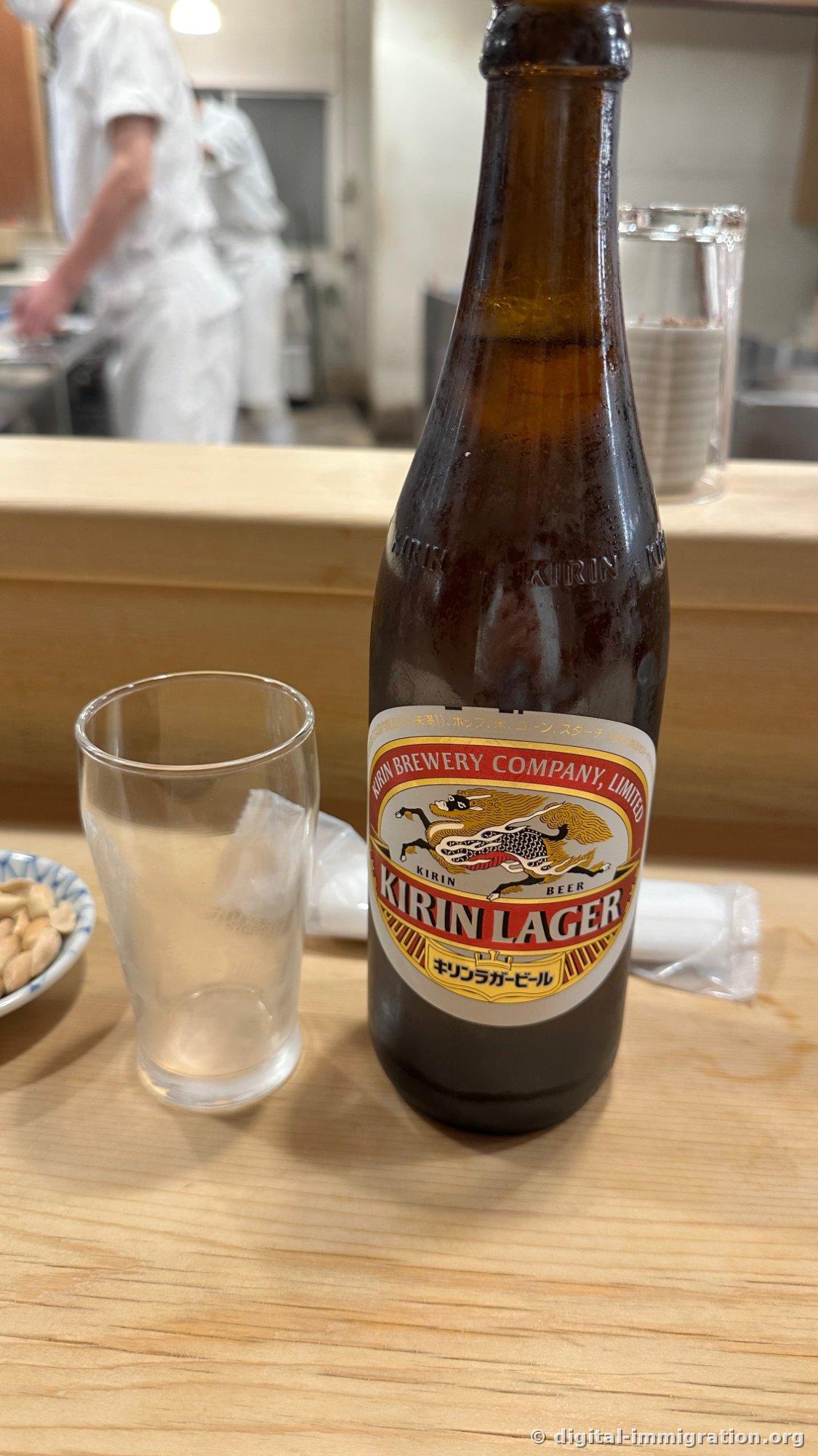 A large bottle of Kirin Lager and an empty glass on the hinoki counter, with kitchen staff in white chef's coats working at the stove behind, stacked water glasses visible to the side