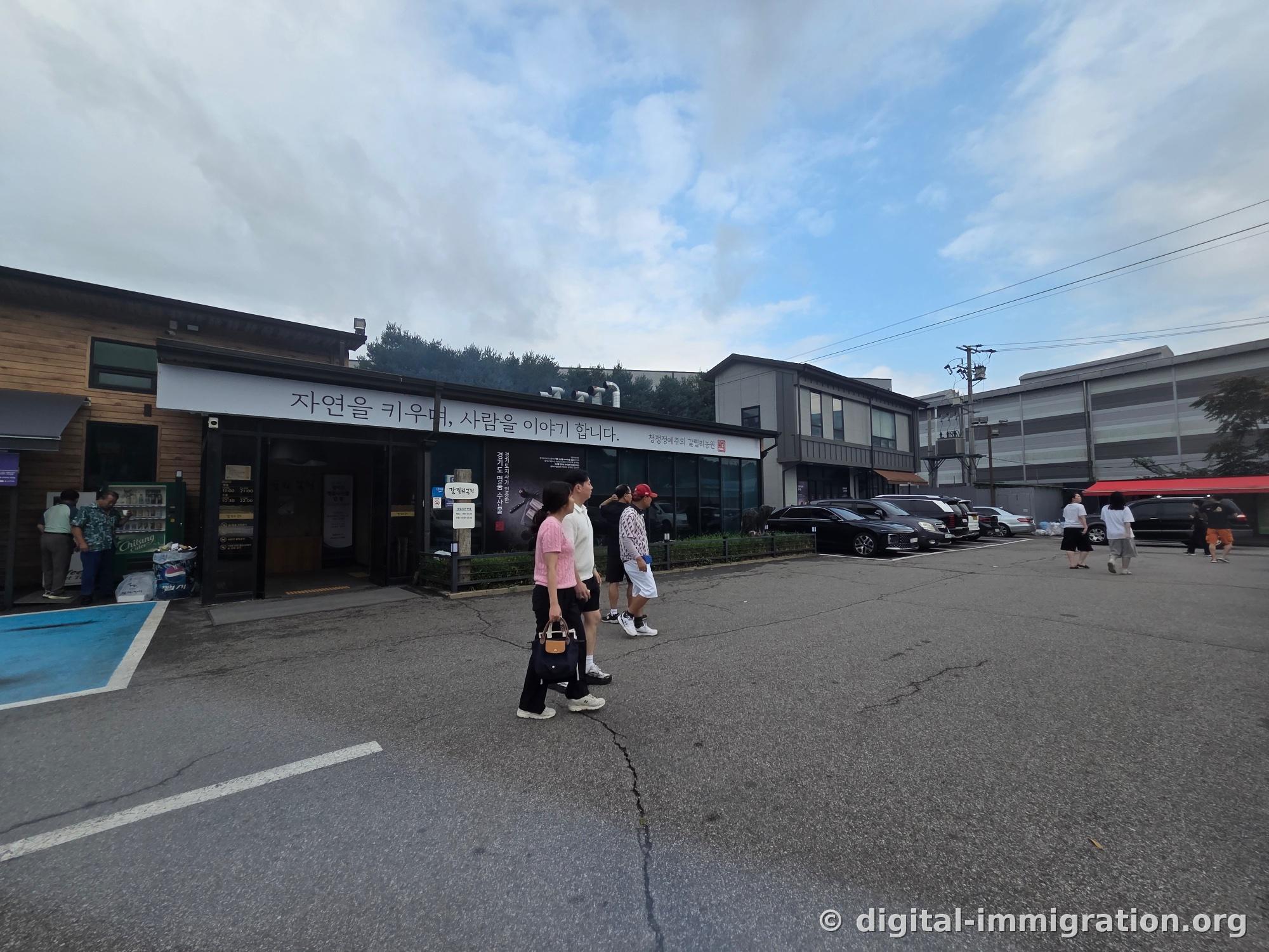 Sous un ciel couvert, la façade de la Ferme Galilée avec l'inscription « Nous cultivons la nature et racontons les histoires des gens », le vaste parking et les clients qui arrivent
