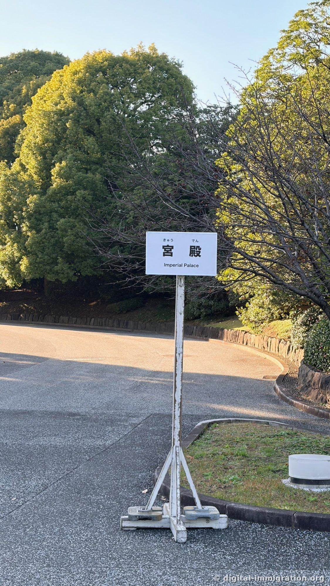 Un panneau blanc au bord de la route indiquant '宮殿 Imperial Palace' avec des arbres verts derrière