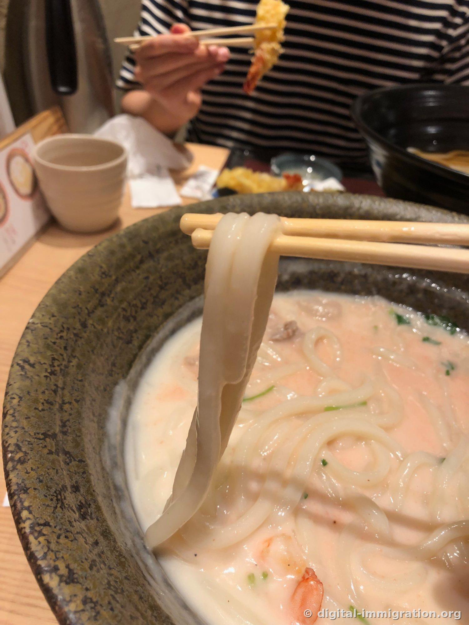 Wide, thick udon noodles lifted with chopsticks, with a companion holding shrimp tempura in the background