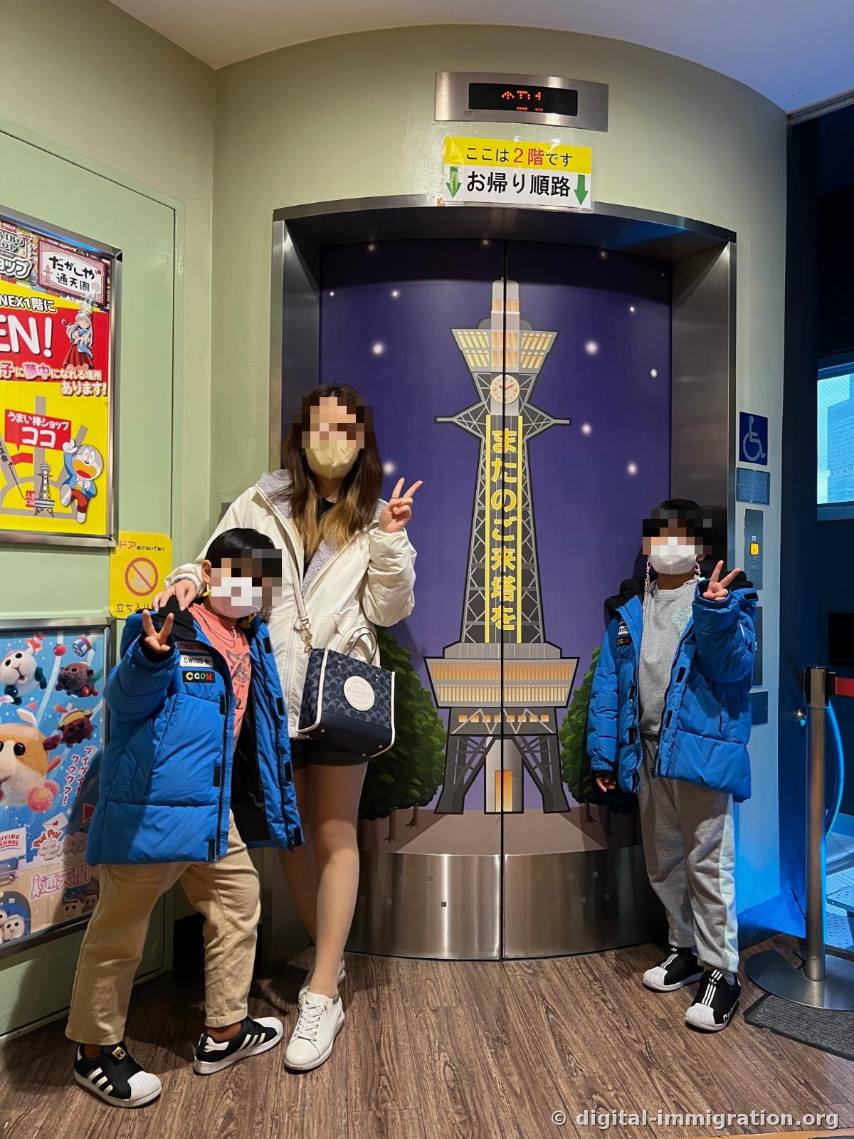 Three people making peace signs in front of an elevator door with a Tsutenkaku illustration